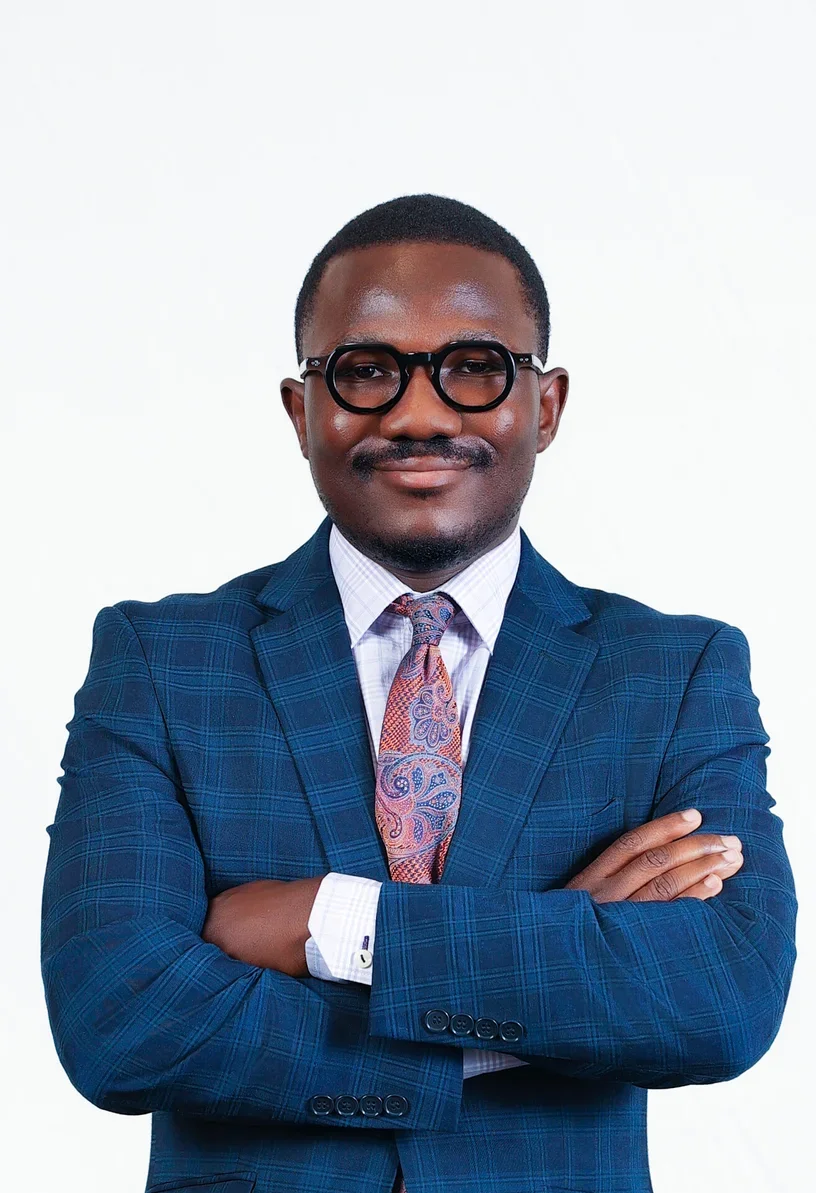 A professional man in a blue checkered suit, white shirt, and colorful paisley tie, wearing round black glasses, standing with arms crossed in front of a plain white background.
