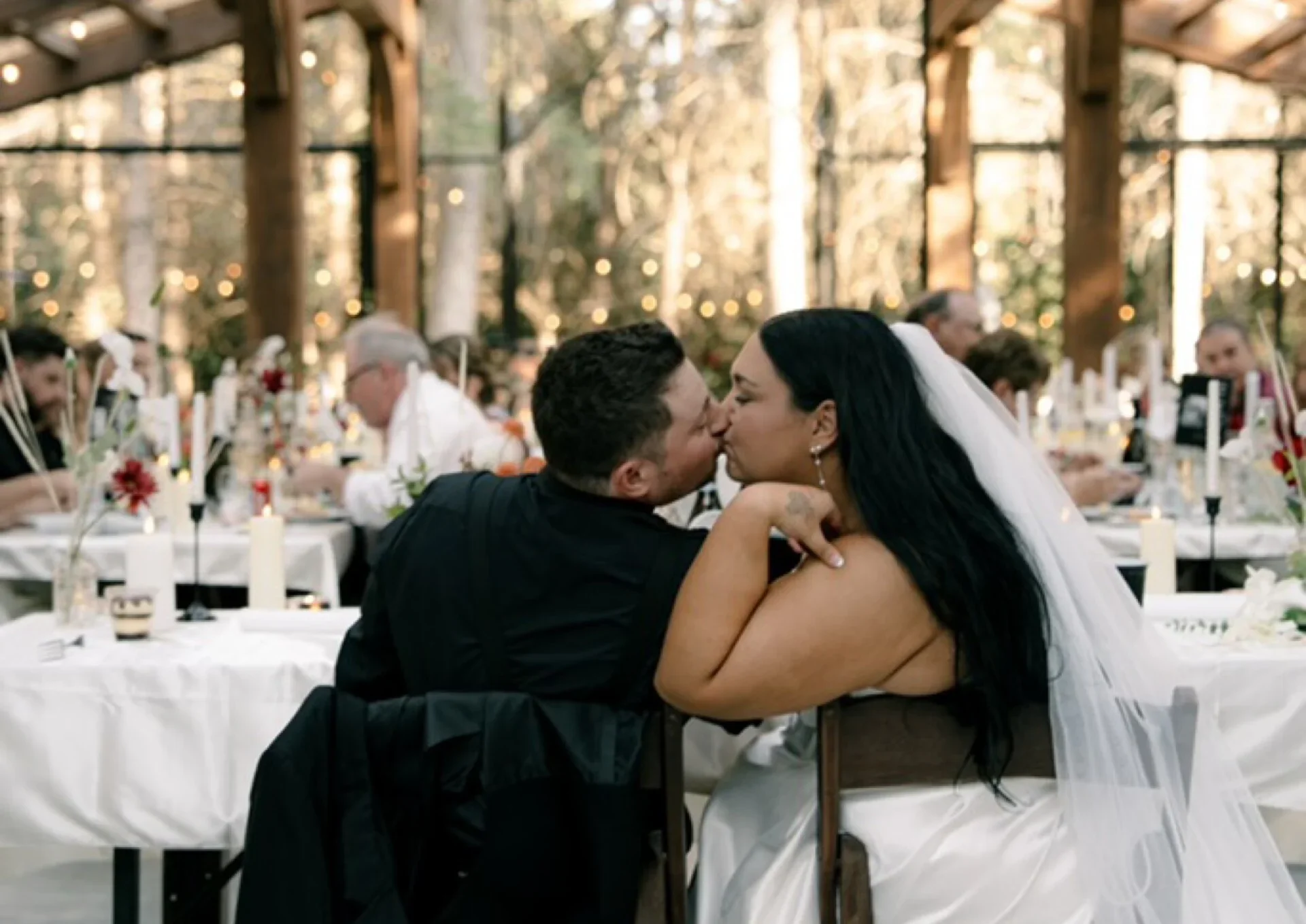 A newlywed couple sharing a kiss at their wedding reception, surrounded by guests and decorated tables in an outdoor setting with trees and string lights.