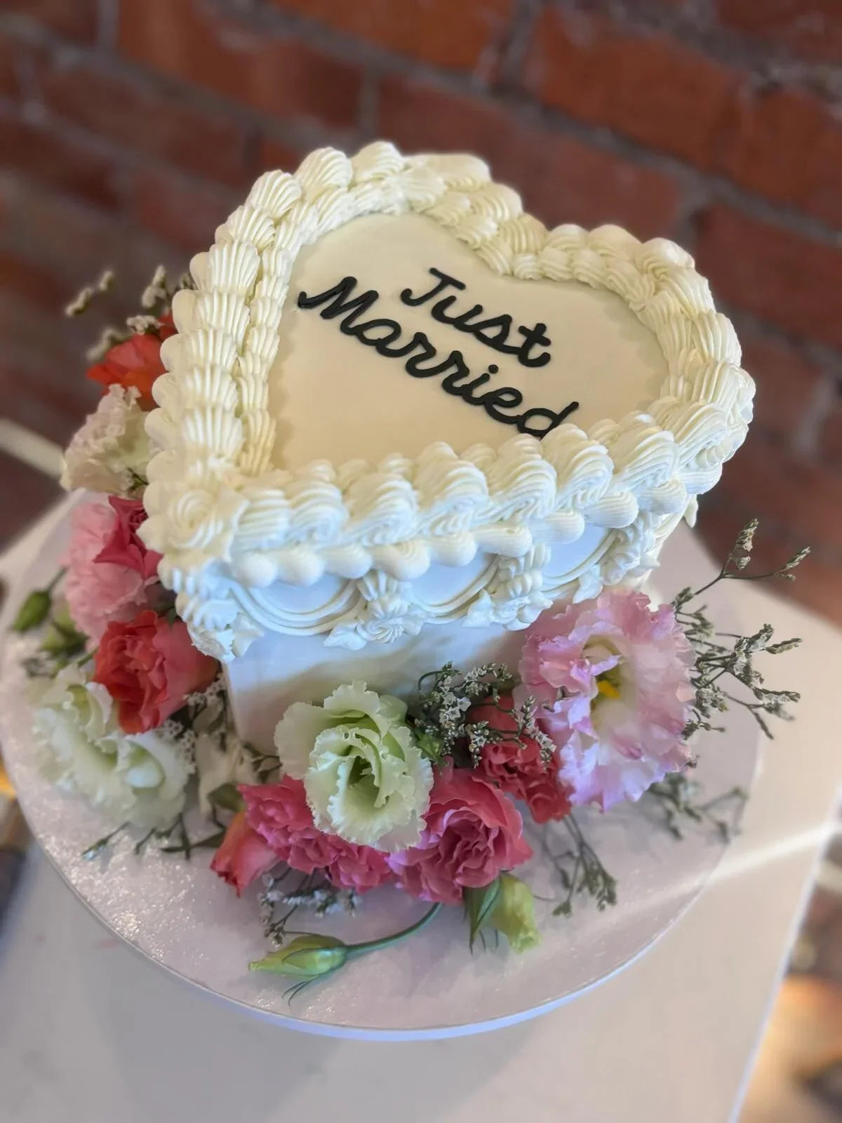 A wedding cake with the message 'Just Married' written on it, decorated with white frosting and surrounded by pink and white flowers, with a brick wall in the background.