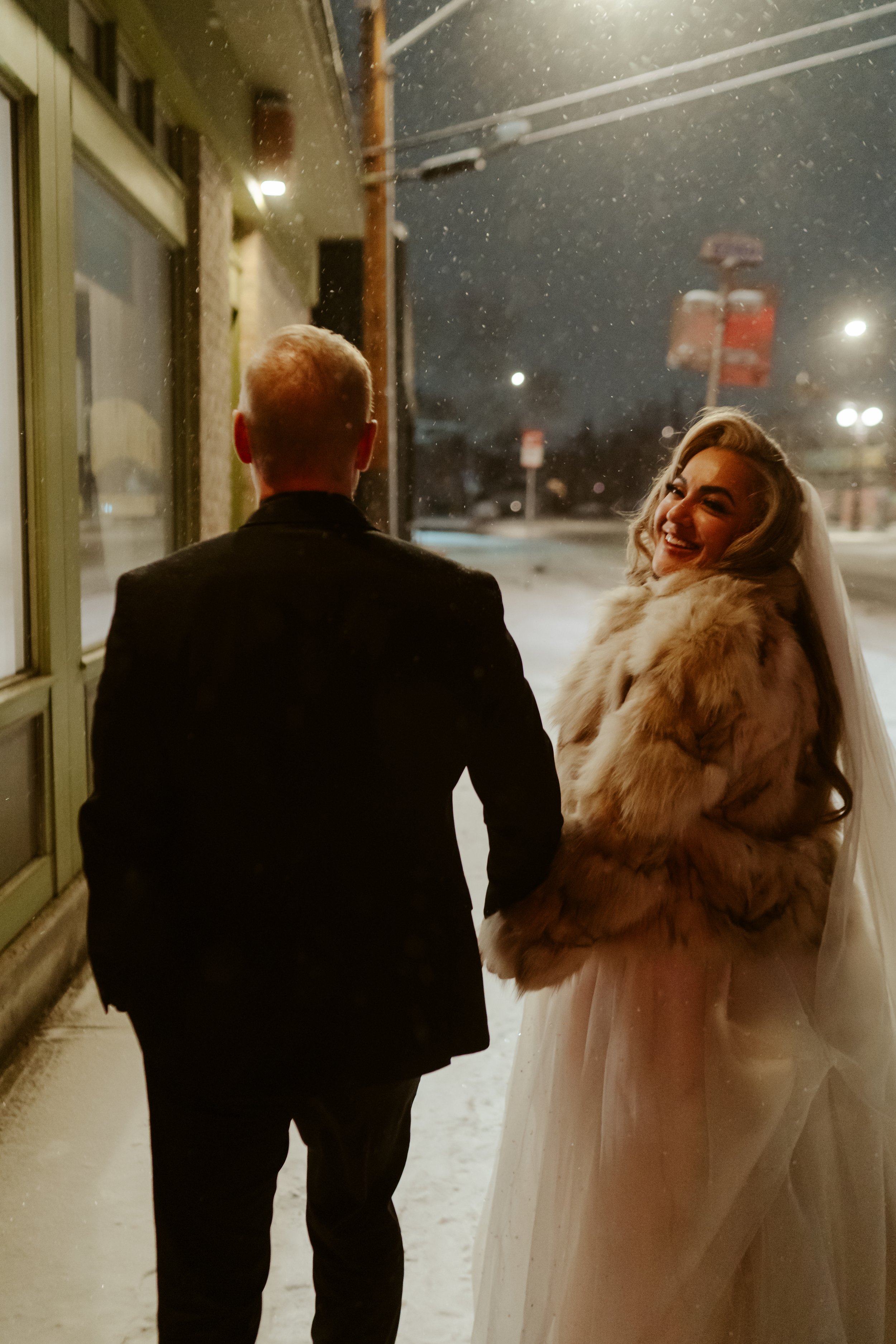 A bride and groom holding hands outside in the snow at night, smiling and looking at each other.