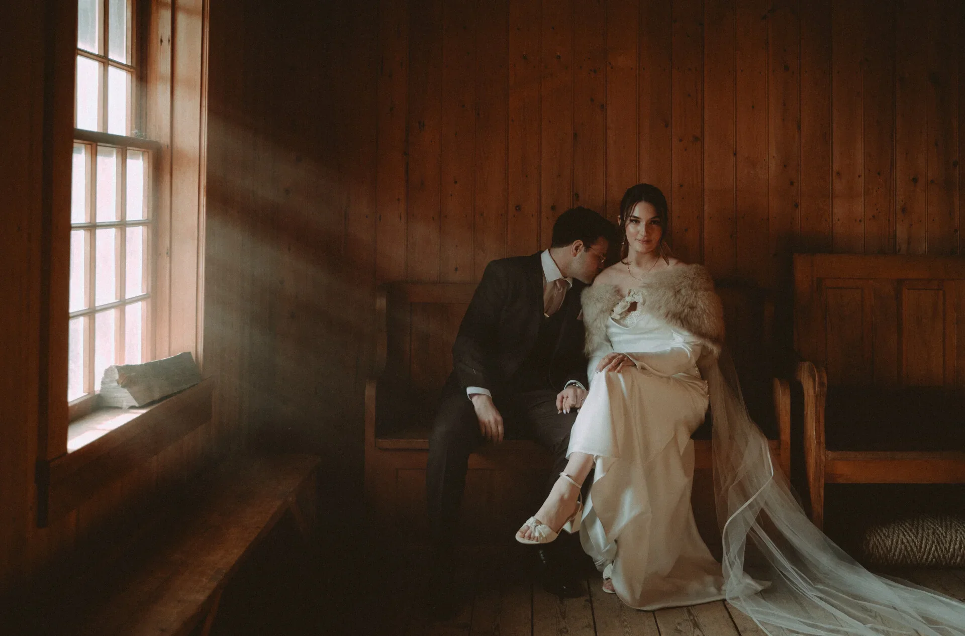 A bride and groom sitting on a wooden bench in a warmly lit room with wood-paneled walls, the bride in a white gown with a fur wrap and sheer veil, the groom in a dark suit leaning in to kiss the bride's cheek.
