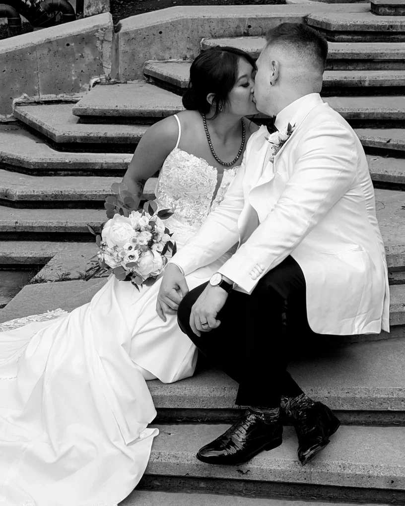 A black and white photo of a bride and groom sharing a kiss, sitting on outdoor steps. The bride wears a lace wedding dress and holds a bouquet, while the groom wears a tuxedo and a white jacket.