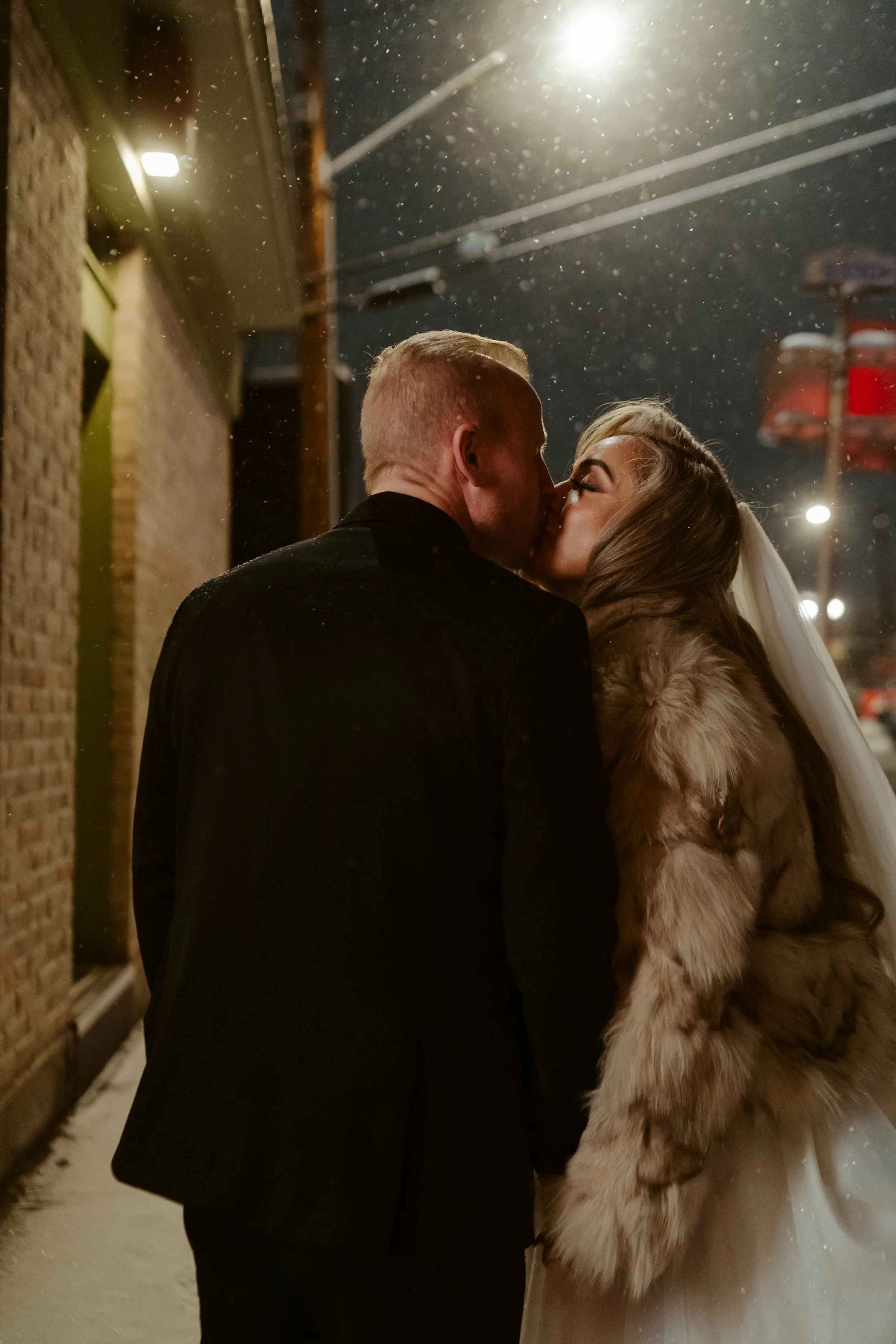 A couple in wedding attire sharing a kiss outside at night during snowfall, with streetlights and utility poles visible in the background.