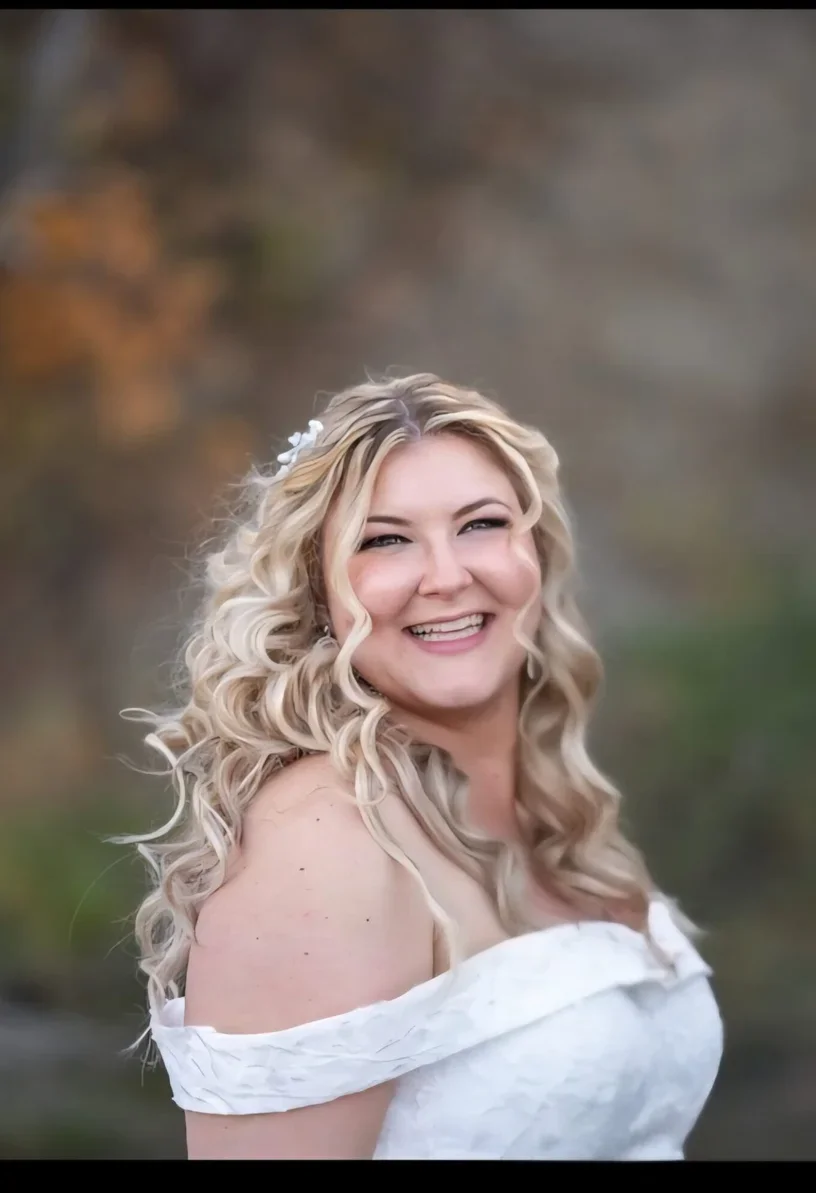 A woman with long, curly blonde hair smiling outdoors with autumn trees in the background.