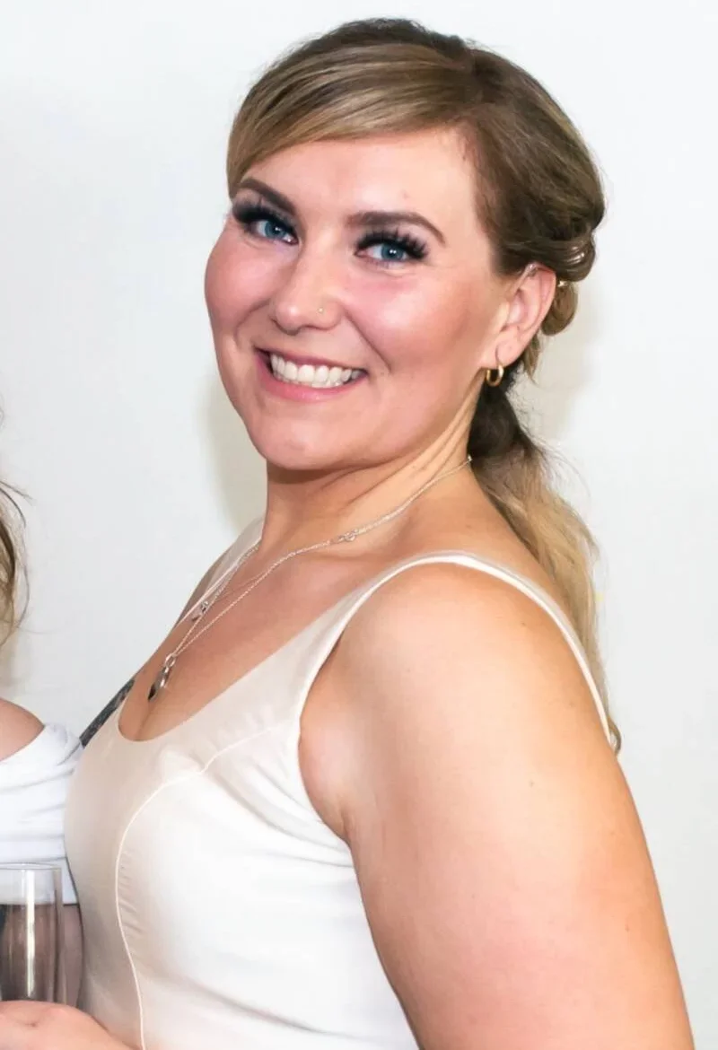 A woman with blonde hair styled to the side, wearing a white satin dress, gold hoop earrings, a delicate necklace, and smiling with blue eyes and makeup.