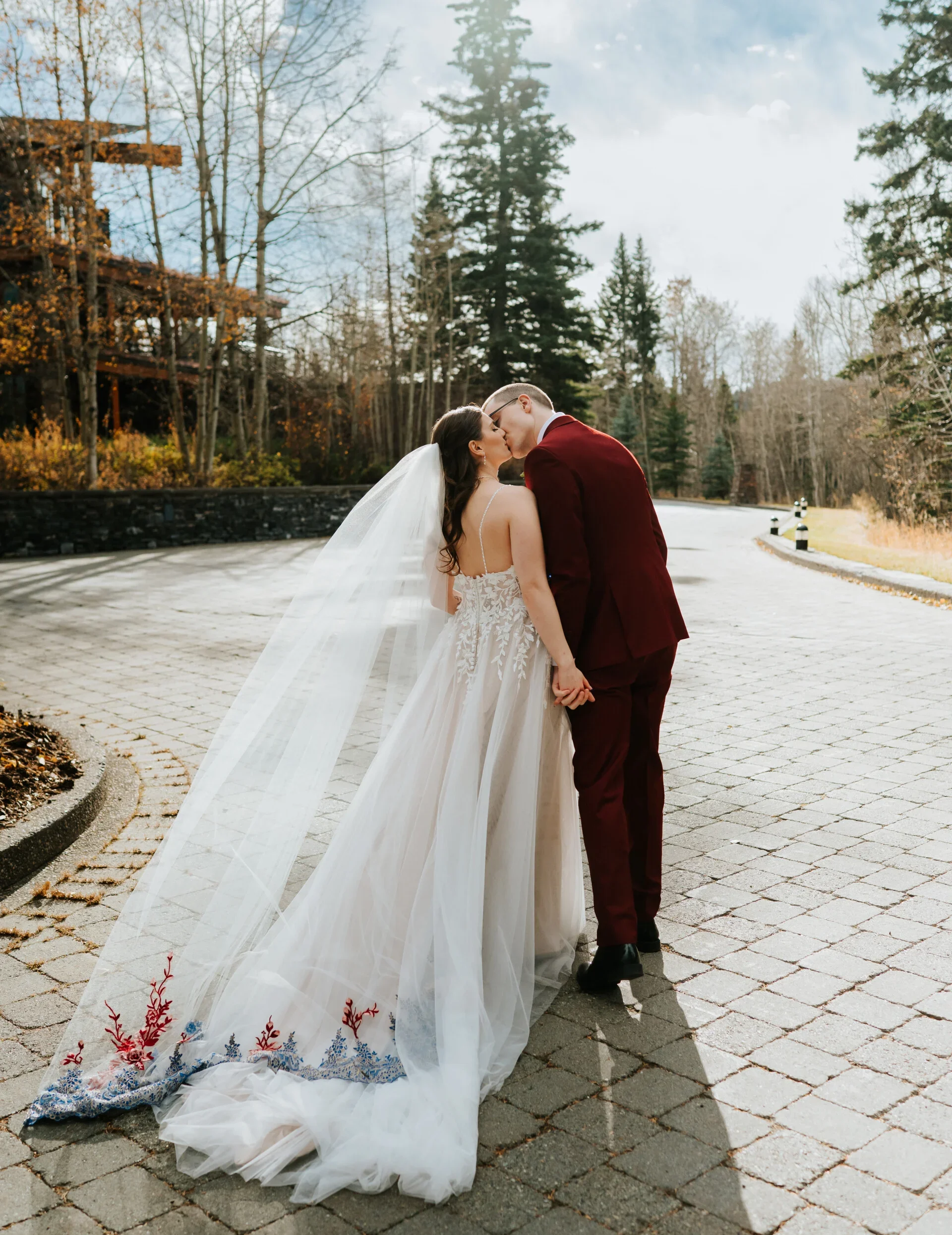 A bride and groom kissing outdoors on a paved road, holding hands, with trees in the background on a cloudy day.