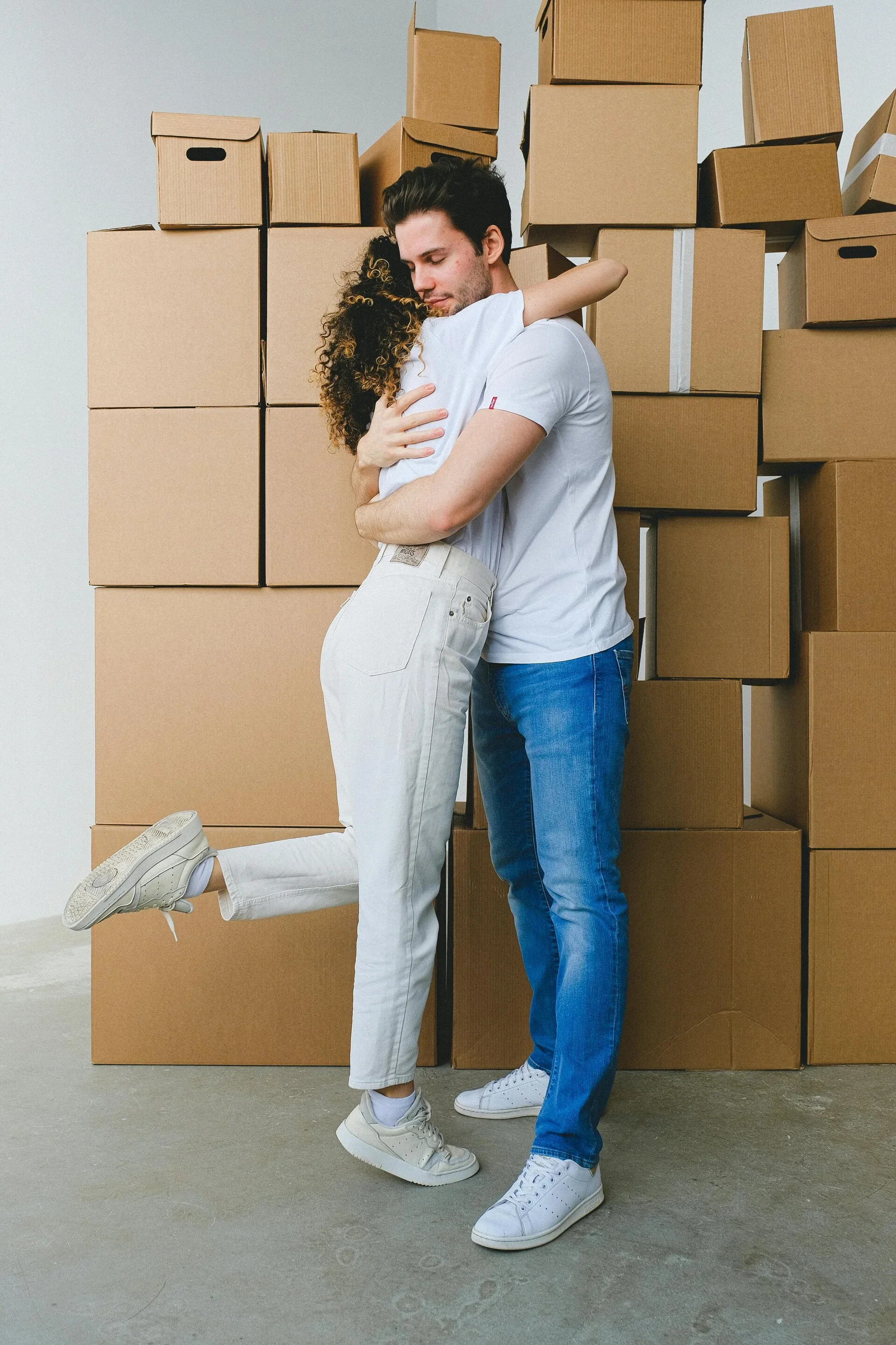 A couple hugging in front of stacked cardboard boxes, with the woman lifting her leg and wrapping her arms around the man's neck.
