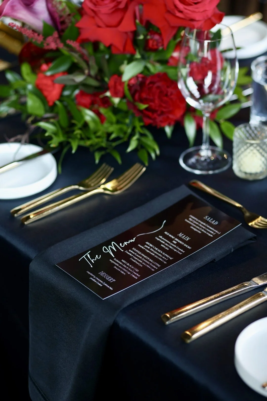 A custom menu card on a formal table setting with a black tablecloth, gold utensils, a wine glass, a small stone plate, and a floral centerpiece of red roses.