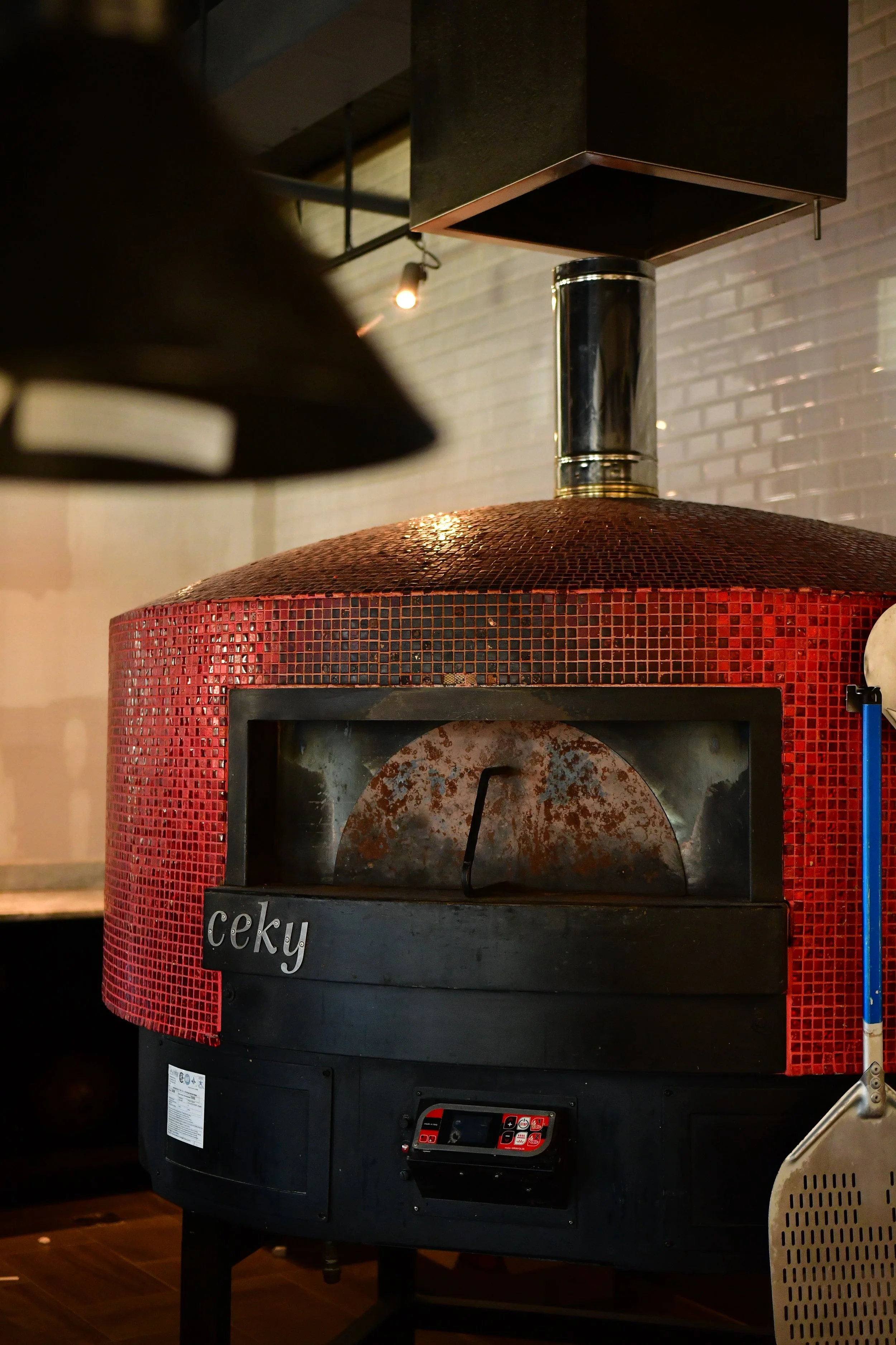 A red mosaic-style wood fired pizza oven with a black door and a metal chimney on top. The oven has a digital control panel and a label that says 'ceky.'
