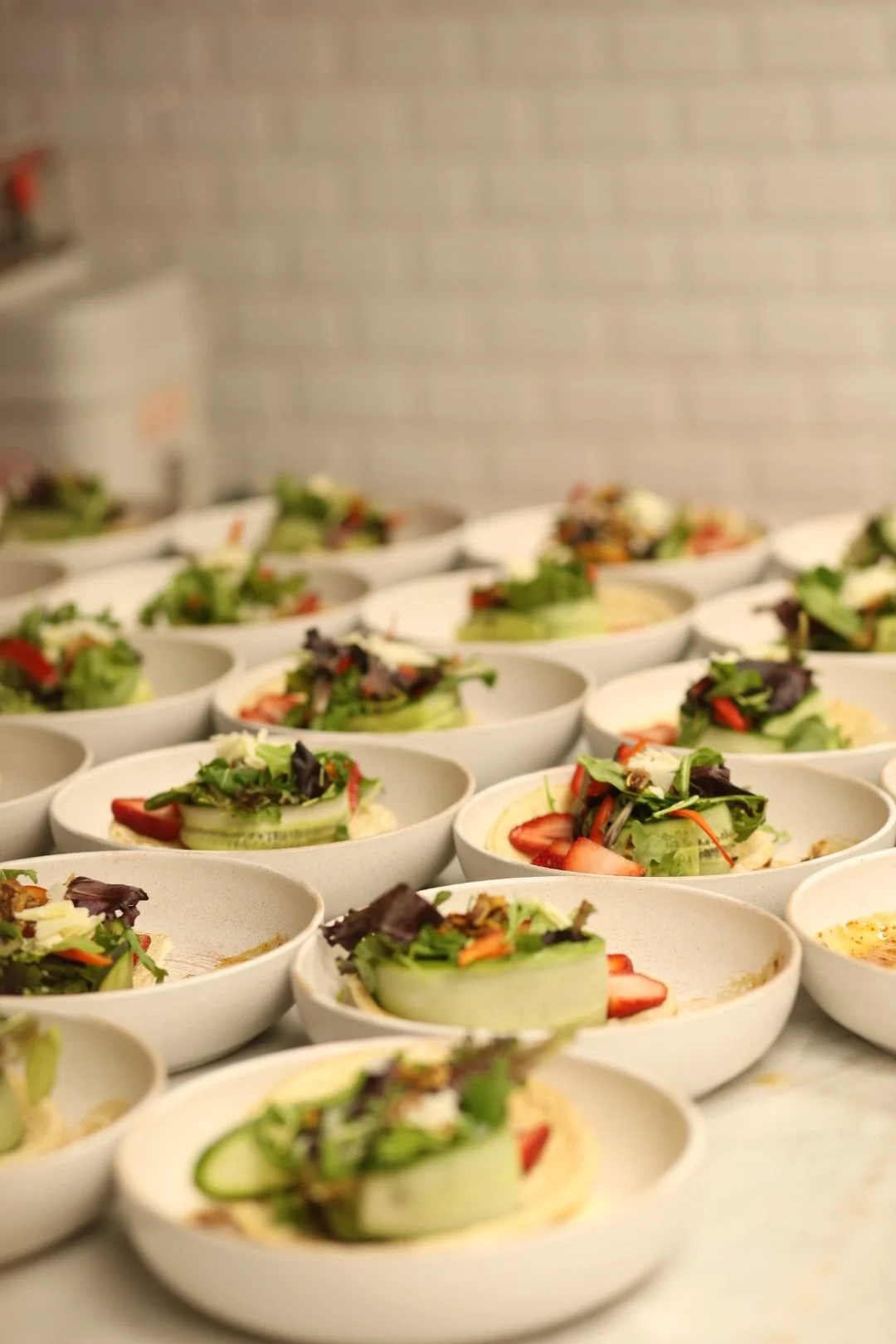 Multiple bowls of fresh salad with mixed greens, strawberries, cucumber slices, and shredded cheese on a catering kitchen countertop.