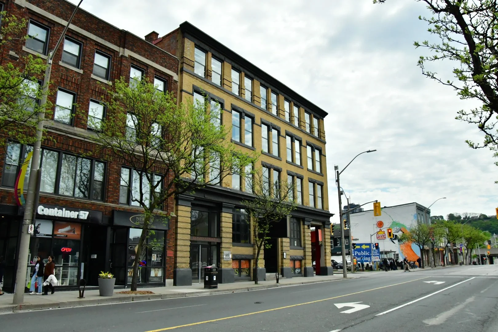 Street view of a city block with mixed-use buildings, storefronts, trees, and signage. People are seen walking on the sidewalk under an overcast sky.