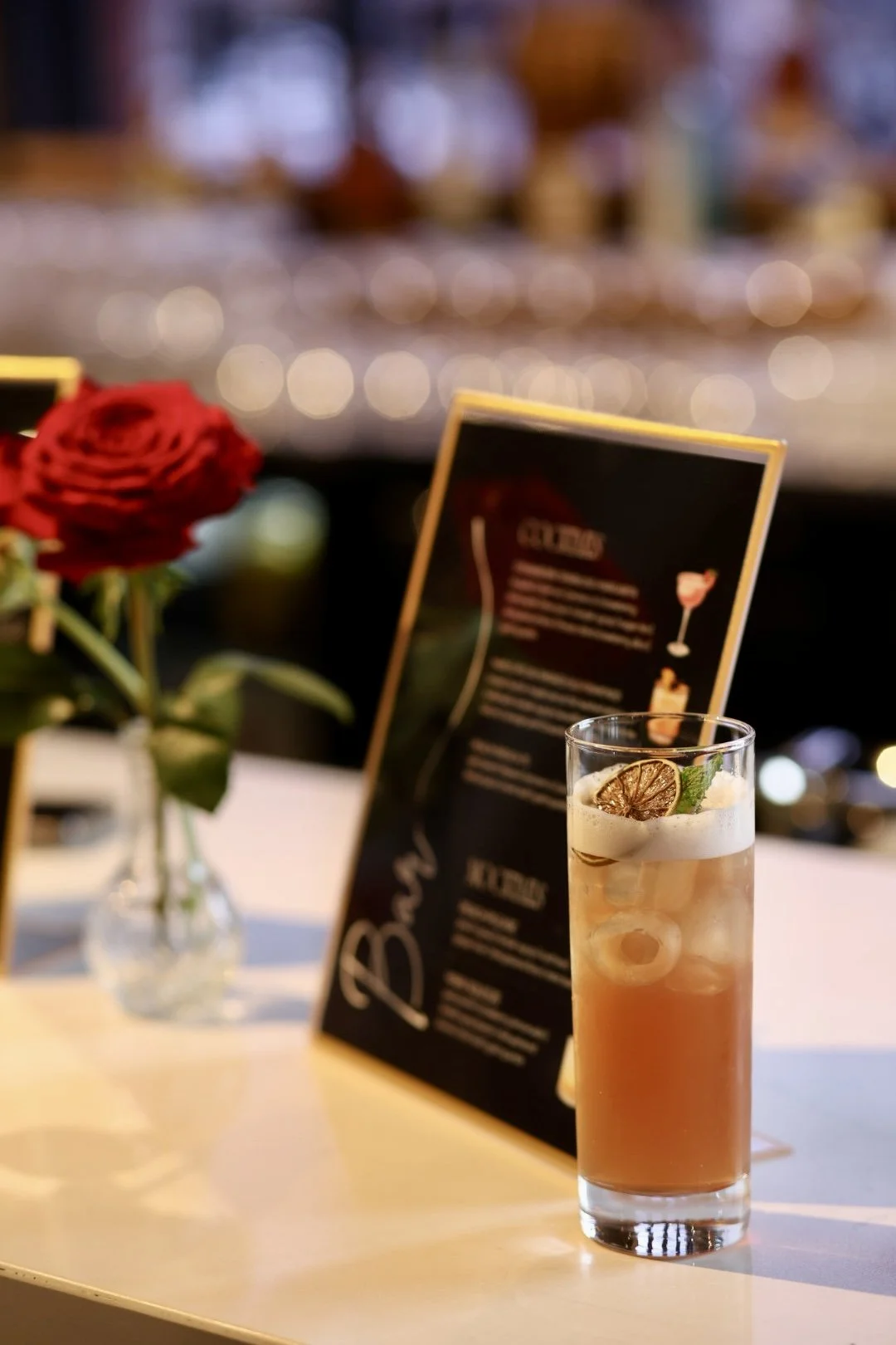 A tall highball glass with a custom cocktail garnished with a slice of dried lime and a sprig of mint, placed on a table next to a small bud vase with a red rose and a bar menu card in the background.