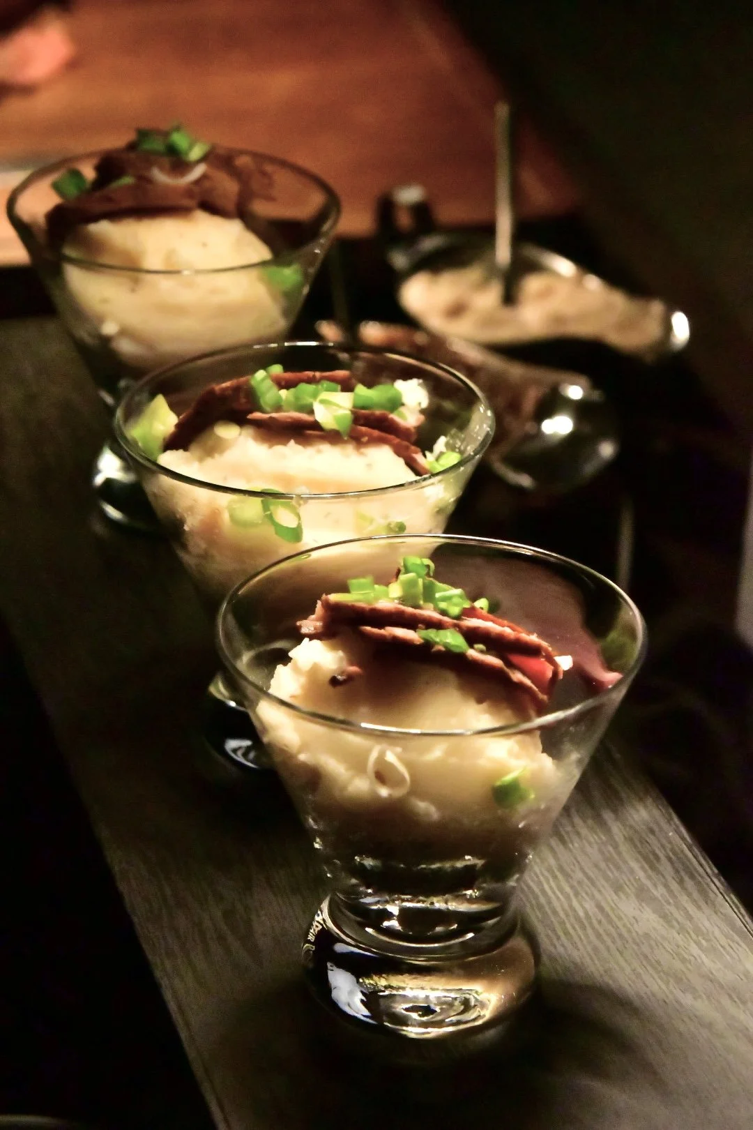 Three martini glasses filled with mashed potatoes topped with carved prime rib and chopped green onions, presented on a wooden serving board.