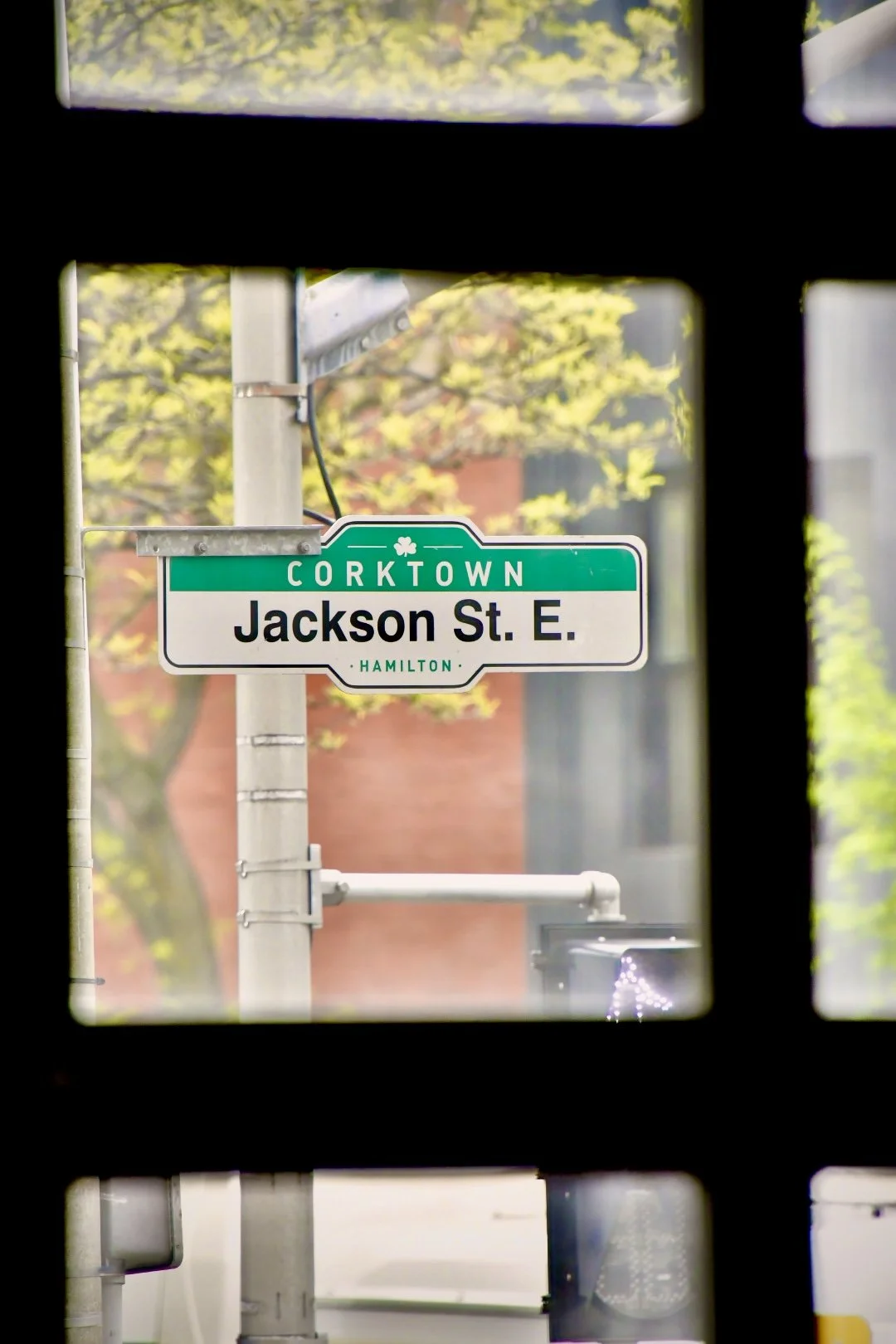 Street sign for Jackson Street East in Corktown neighborhood, Hamilton, Ontario, viewed through a window with black framing, with trees and a brick building in the background.