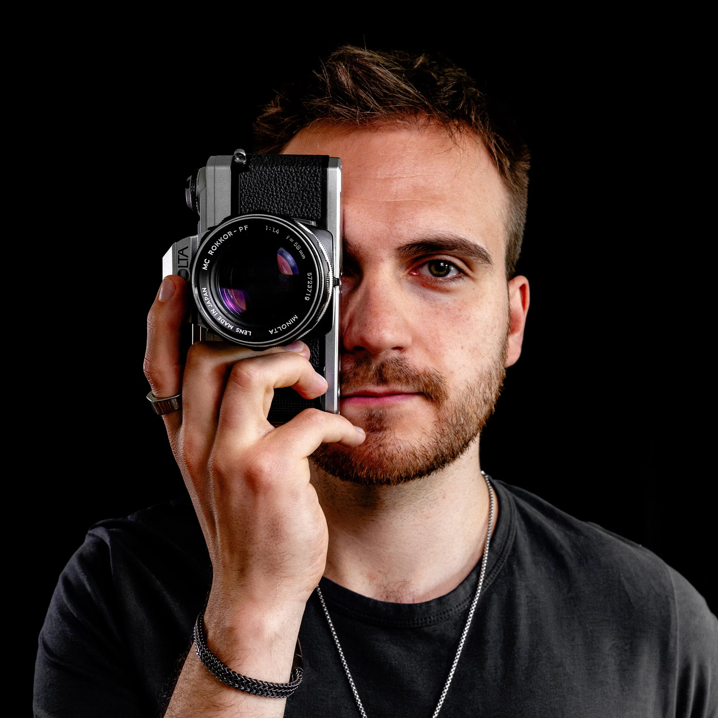 A man with brown hair, a beard, and eye makeup holding a vintage film camera up to his eye, against a black background.