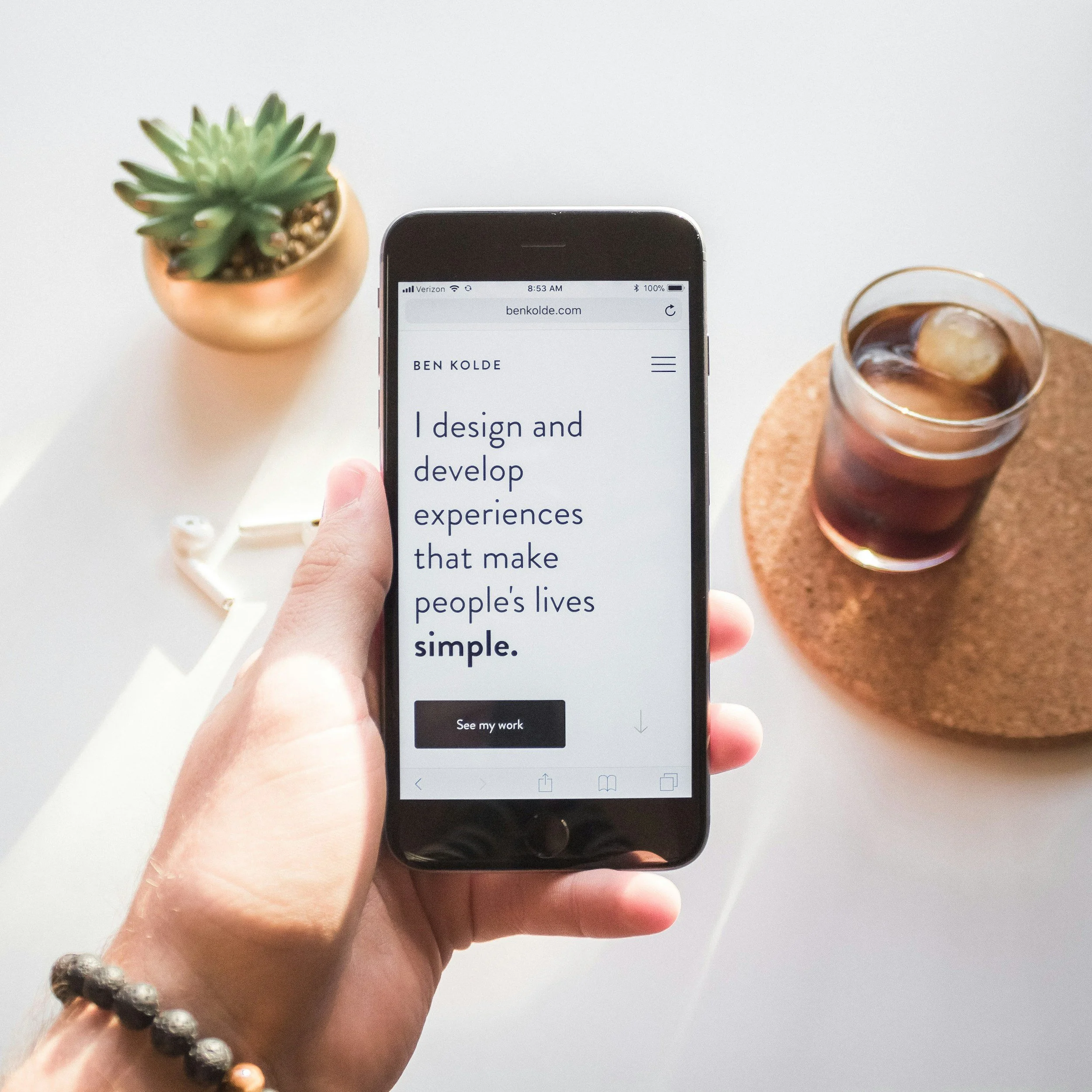 Close-up of a hand holding a smartphone with the website benkolde.com on the screen, and a potted succulent plant and a glass of dark beverage on a cork coaster in the background.