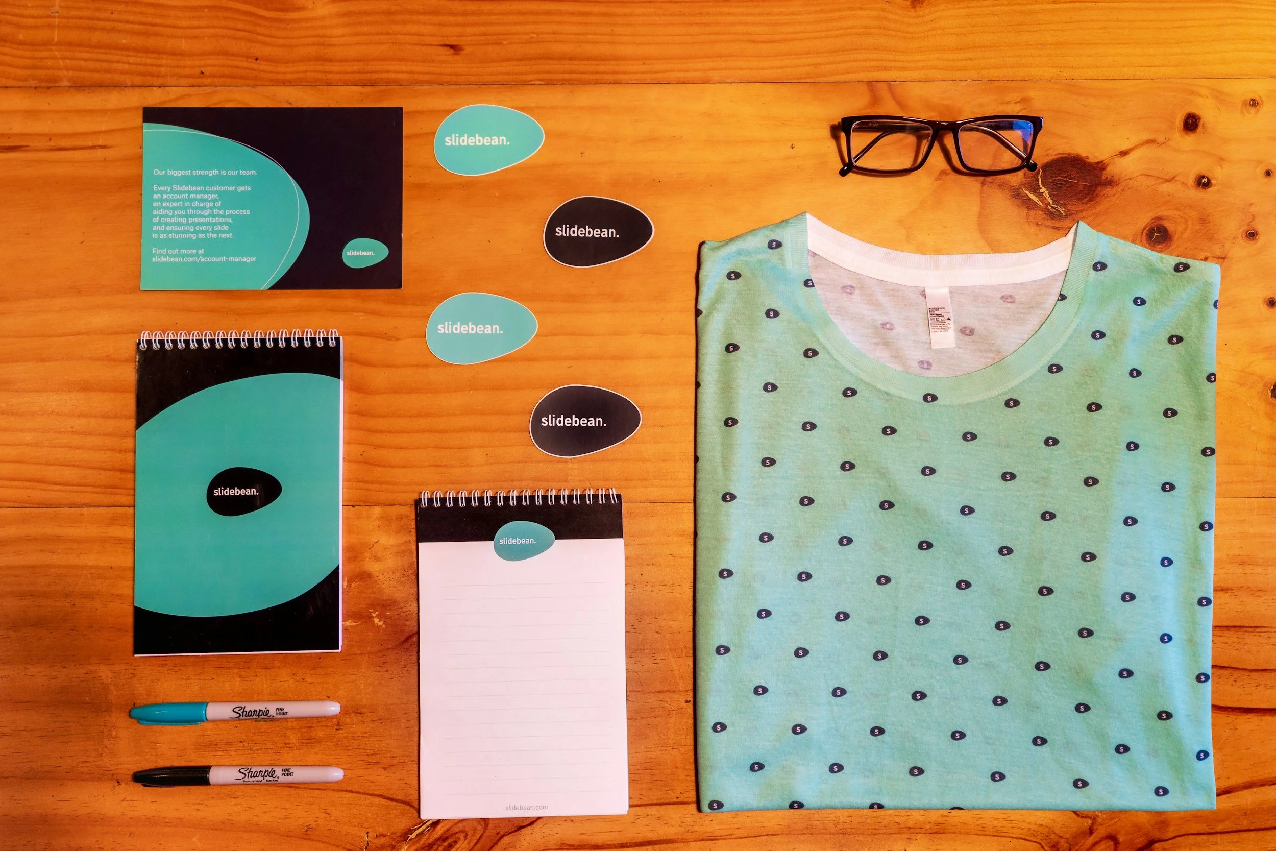 A collection of branded stationery items, including business cards, notebooks, and pens, displayed on a wooden surface. The branding is for Slidebean, featuring a black, teal, and white color scheme with speech bubble designs.