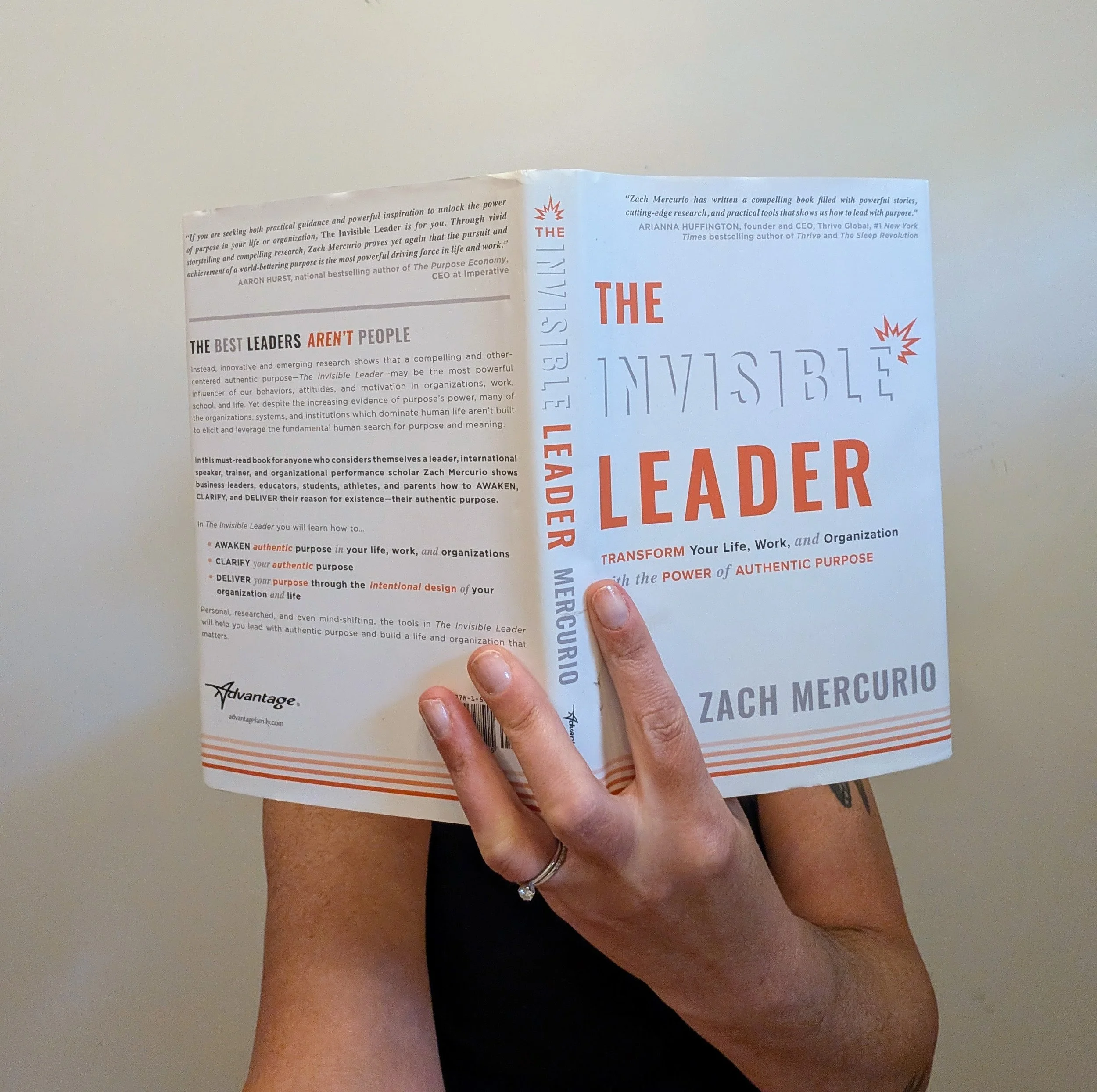 A person holding an open book titled 'The Invisible Leader' by Zach Mercurio. The person's hand and part of their arm are visible, with a ring on their finger. The background is plain and light-colored.