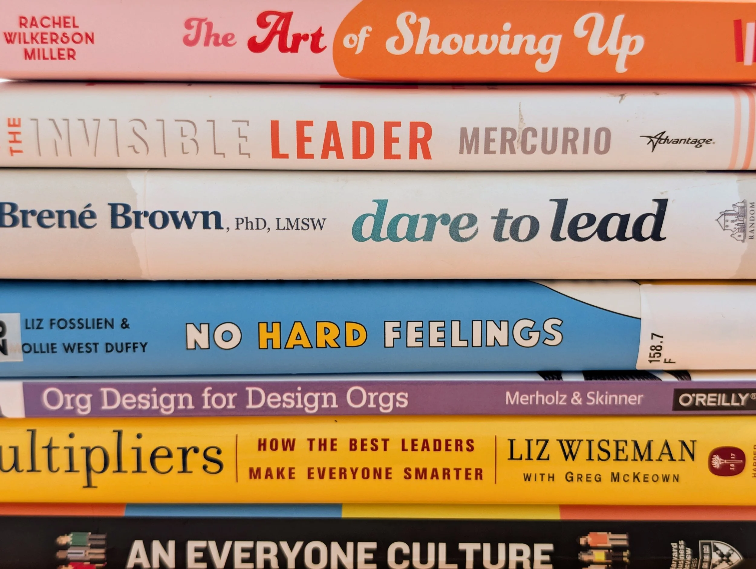 Stack of colorful books with visible titles, including 'The Art of Showing Up', 'The Invisible Leader', 'Dare to Lead', and other titles related to leadership, writing, and culture.