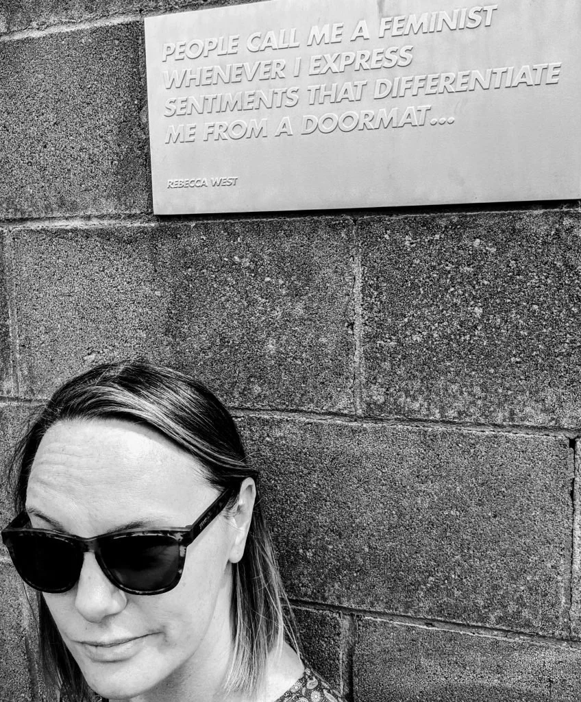 A woman with sunglasses taking a selfie in front of a wall with a quote plaque that reads 'People call me a feminist whenever I express sentiments that differentiate me from a doormat' by Rebecca West.