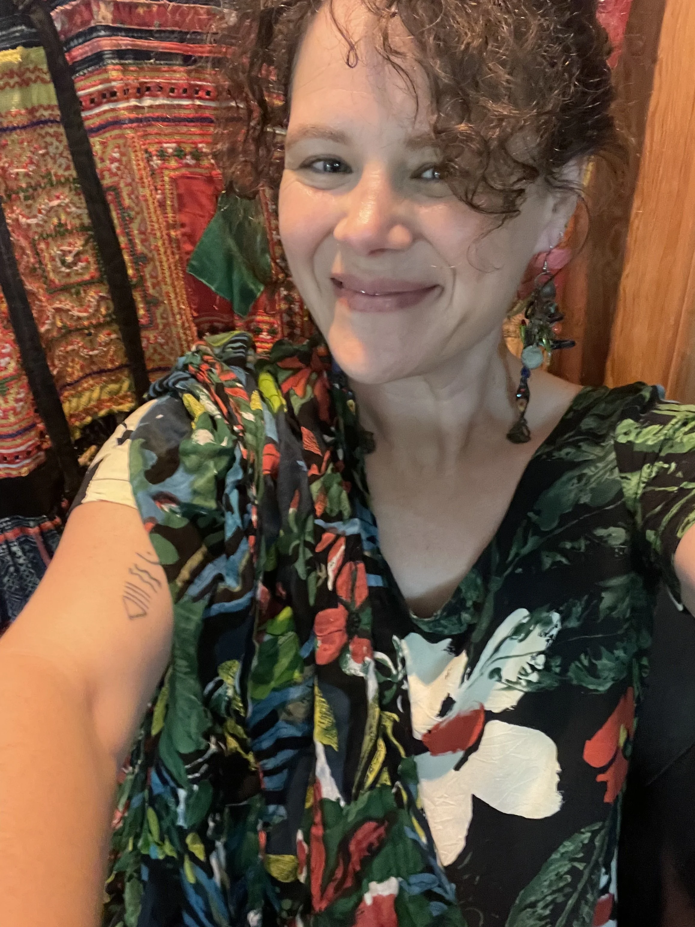 A person taking a selfie with curly hair, wearing a colorful, patterned shirt, standing in front of a vibrant, patterned textile background.