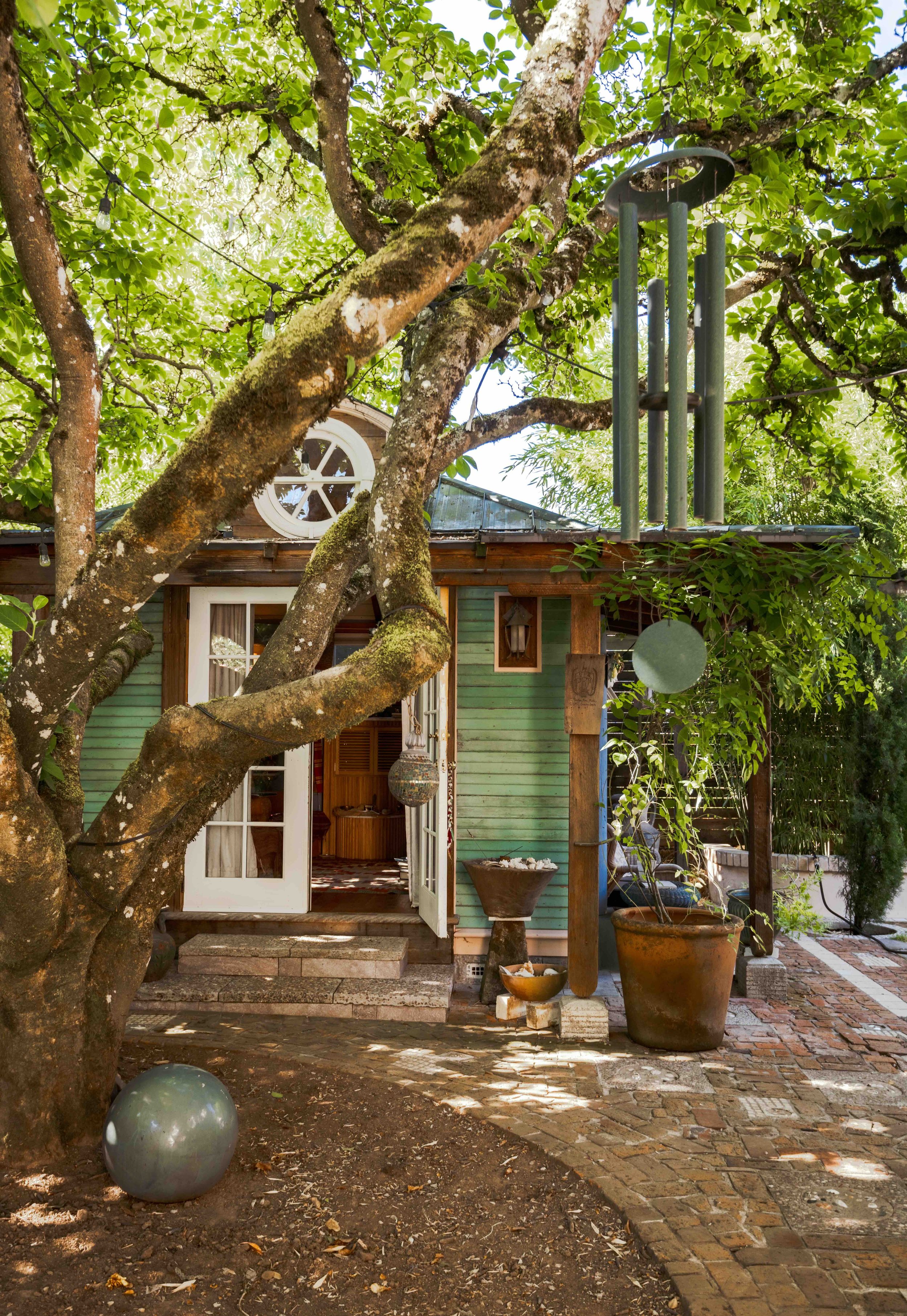 A house surrounded by lush green trees with sunlight filtering through the leaves. The house has a wooden structure with a green exterior and a small porch with steps. There are decorative pots, sculptural objects, and a large tree in the front yard. A wind chime hangs from a tree branch near the house.