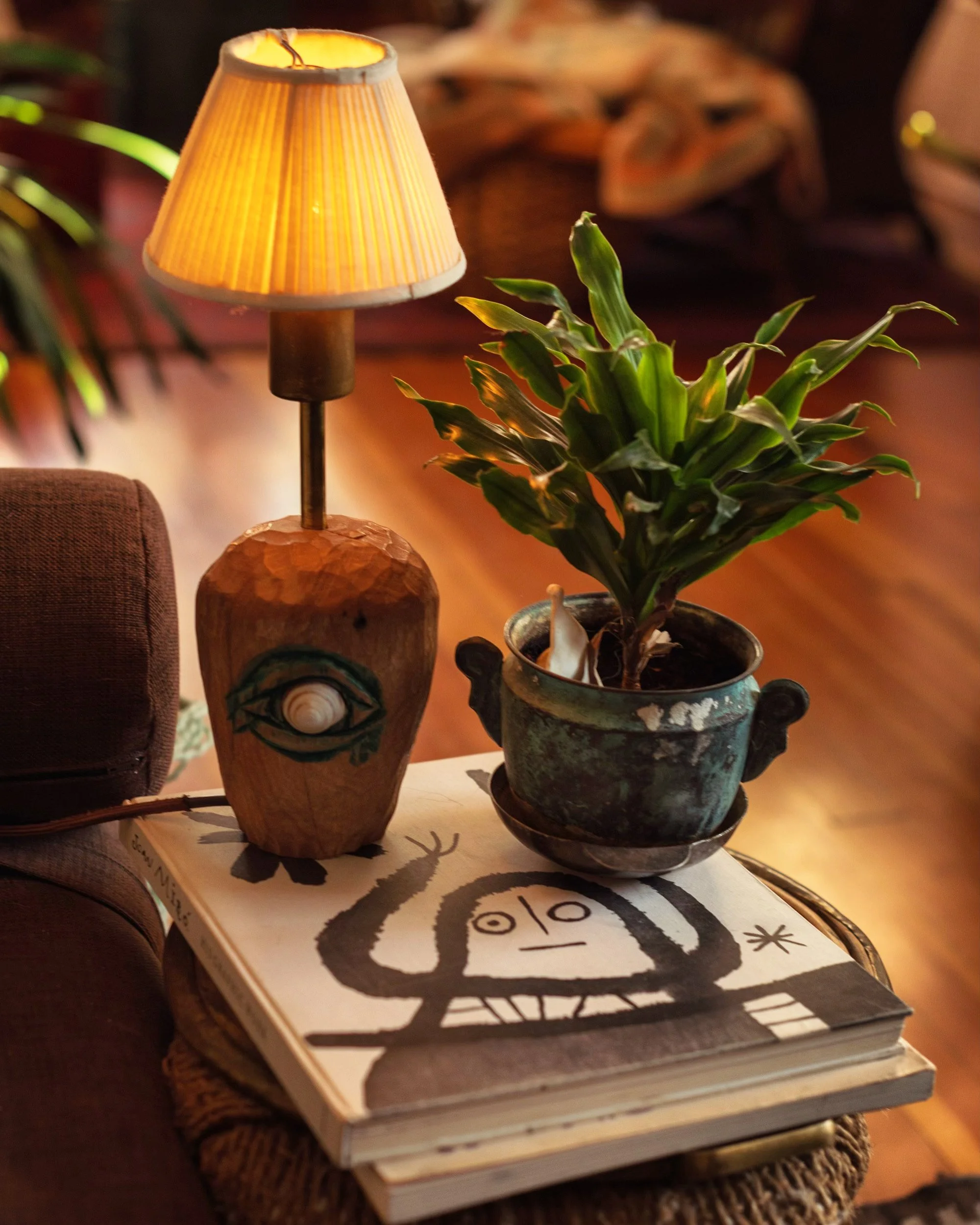 A lit yellow lampshade on a wooden base, a potted green plant, and a stack of illustrated books with a black-and-white drawing of a face and plant on the cover.