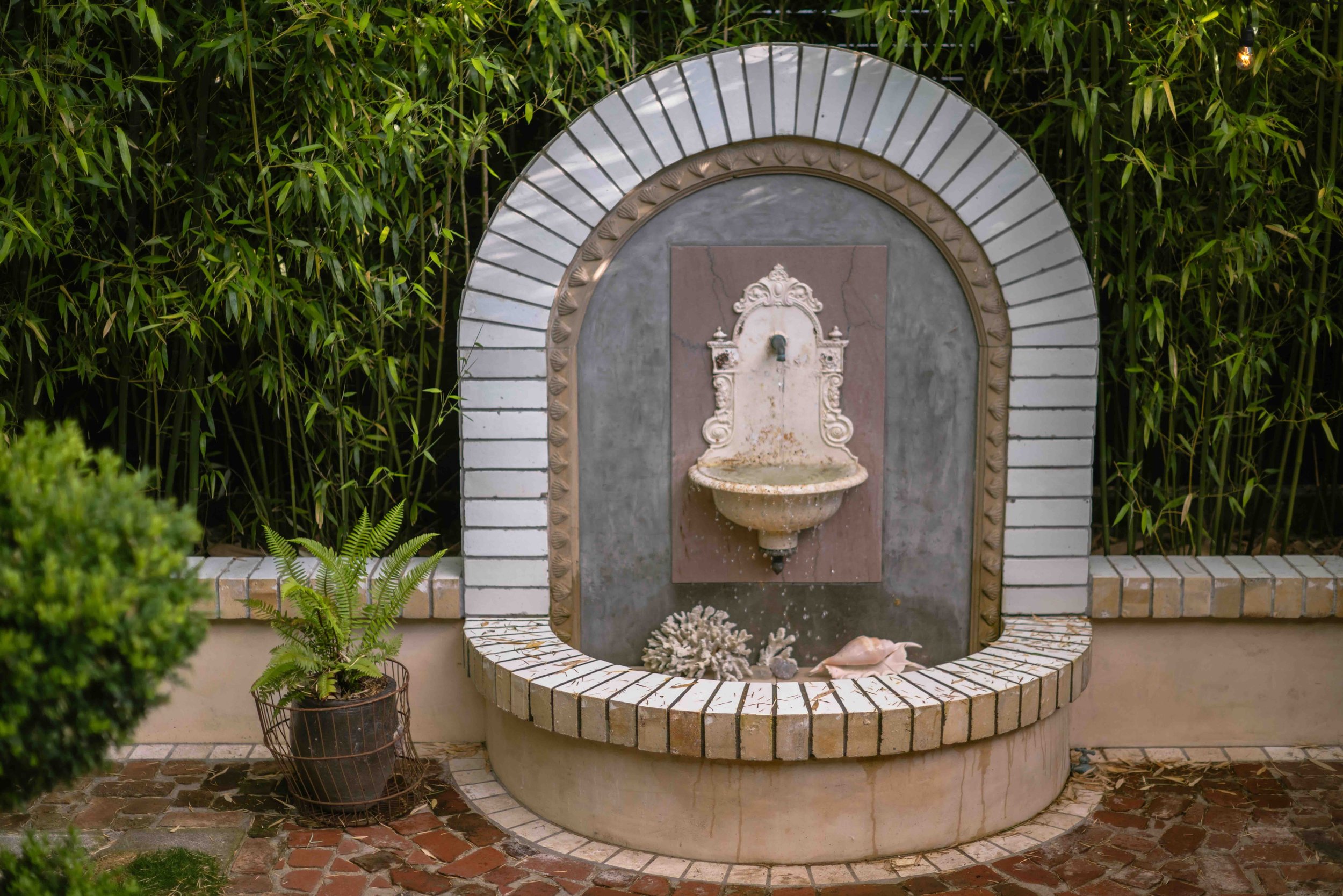 Decorative outdoor fountain with a basin, surrounded by brick and tile, set against a background of green bamboo plants.