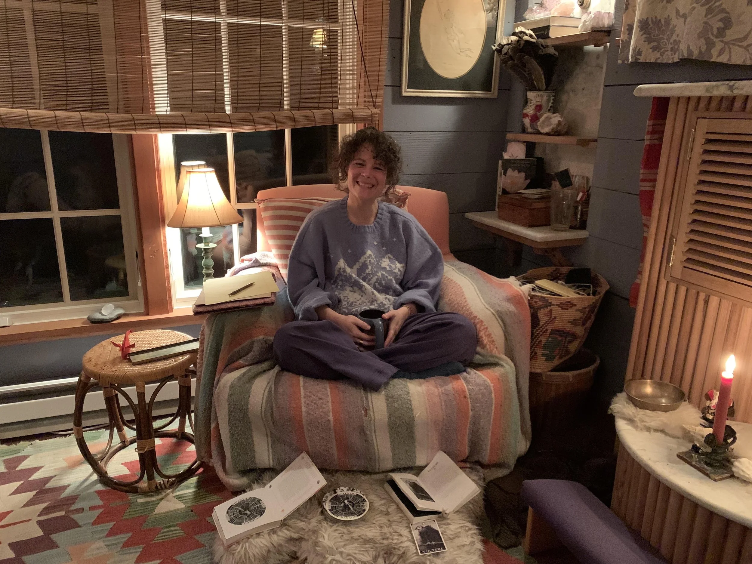 A woman with curly hair and a wide smile sitting on a bed in a cozy room, holding a mug. The room features warm lighting, a lamp, wall art, window blinds, and various decorative items including candles, books, and artwork.