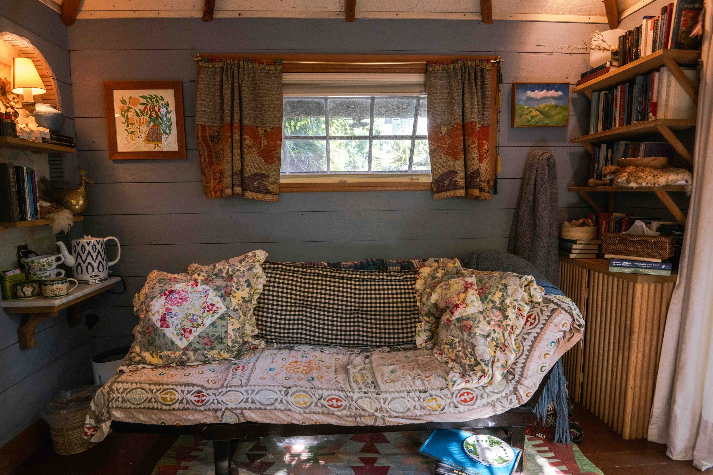 A cozy living room with a large window, a floral sofa, wooden shelves filled with books, and framed artwork on the walls.