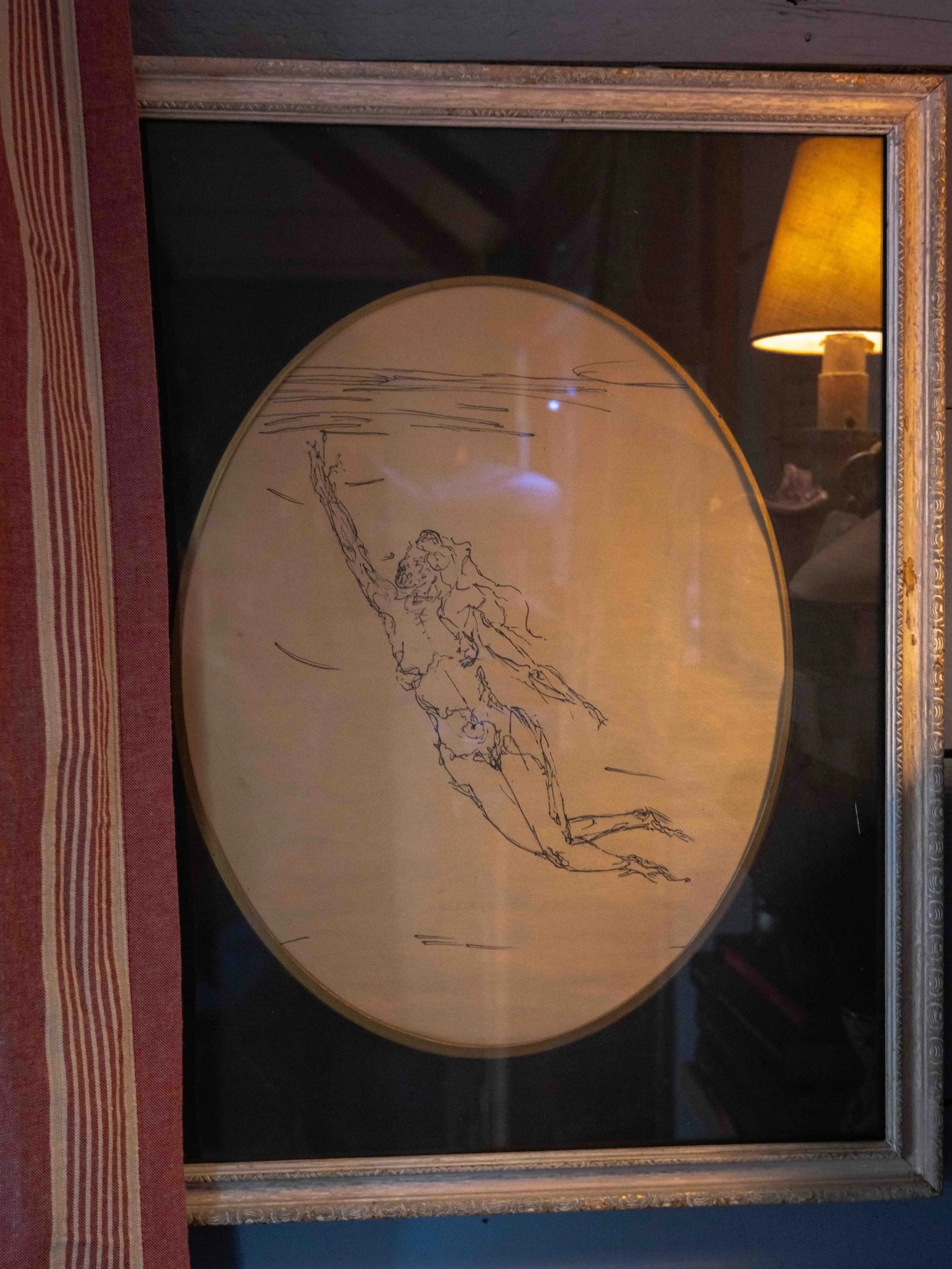 A framed sketch of an angelic figure reaching upwards, with a background resembling water or sky, displayed next to a side table with a lamp.