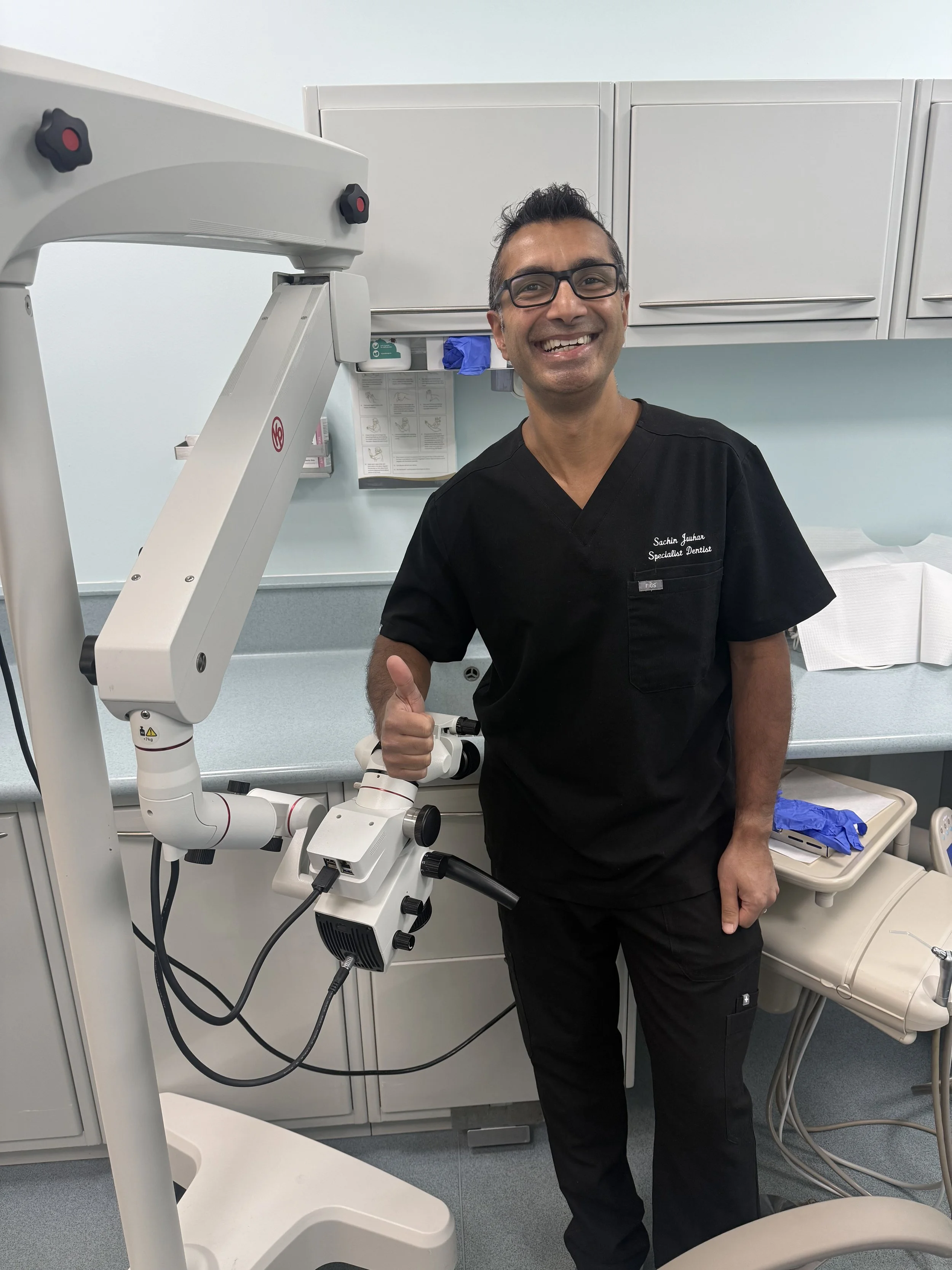 A smiling man in black medical scrubs giving a thumbs-up in a dental office with dental equipment, including a dental microscope, behind him.