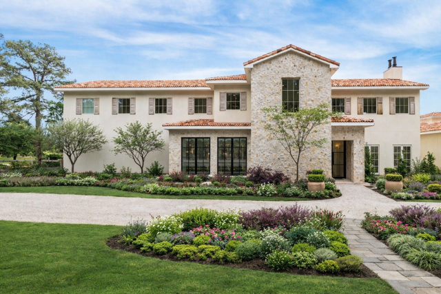 Large house with white stucco walls, a tiled roof, multiple windows, and a stone facade section in the center. Well-maintained garden with colorful flowers and trees, a paved pathway, and a green lawn.