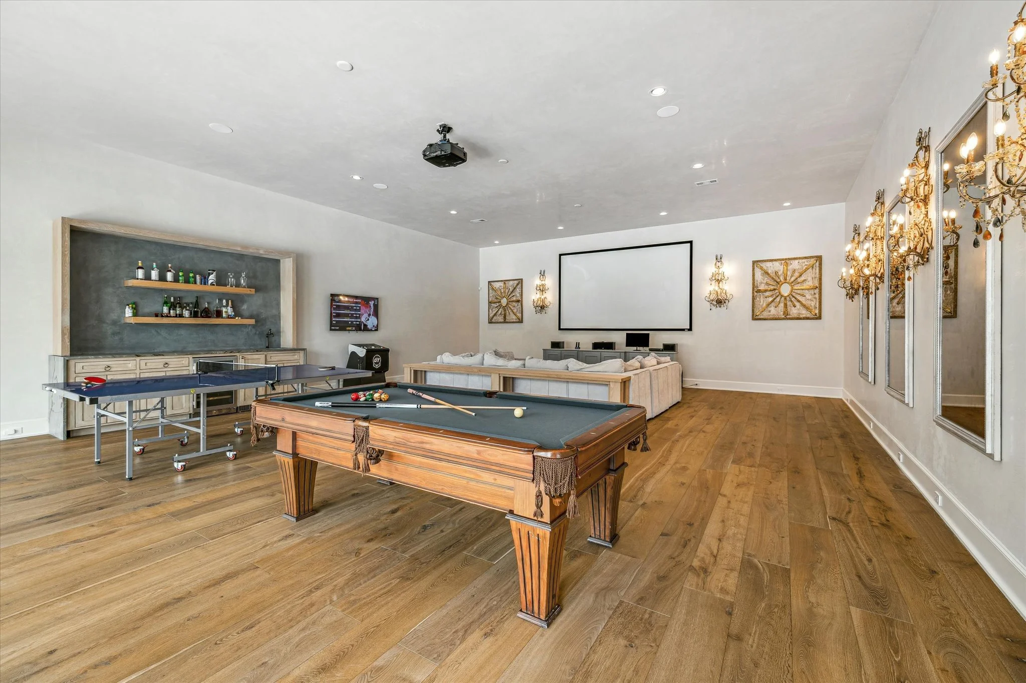 A spacious recreation room with a pool table, ping pong table, and large TV. The room has hardwood floors, a sectional sofa facing a large screen, and decorative chandeliers and wall mirrors.