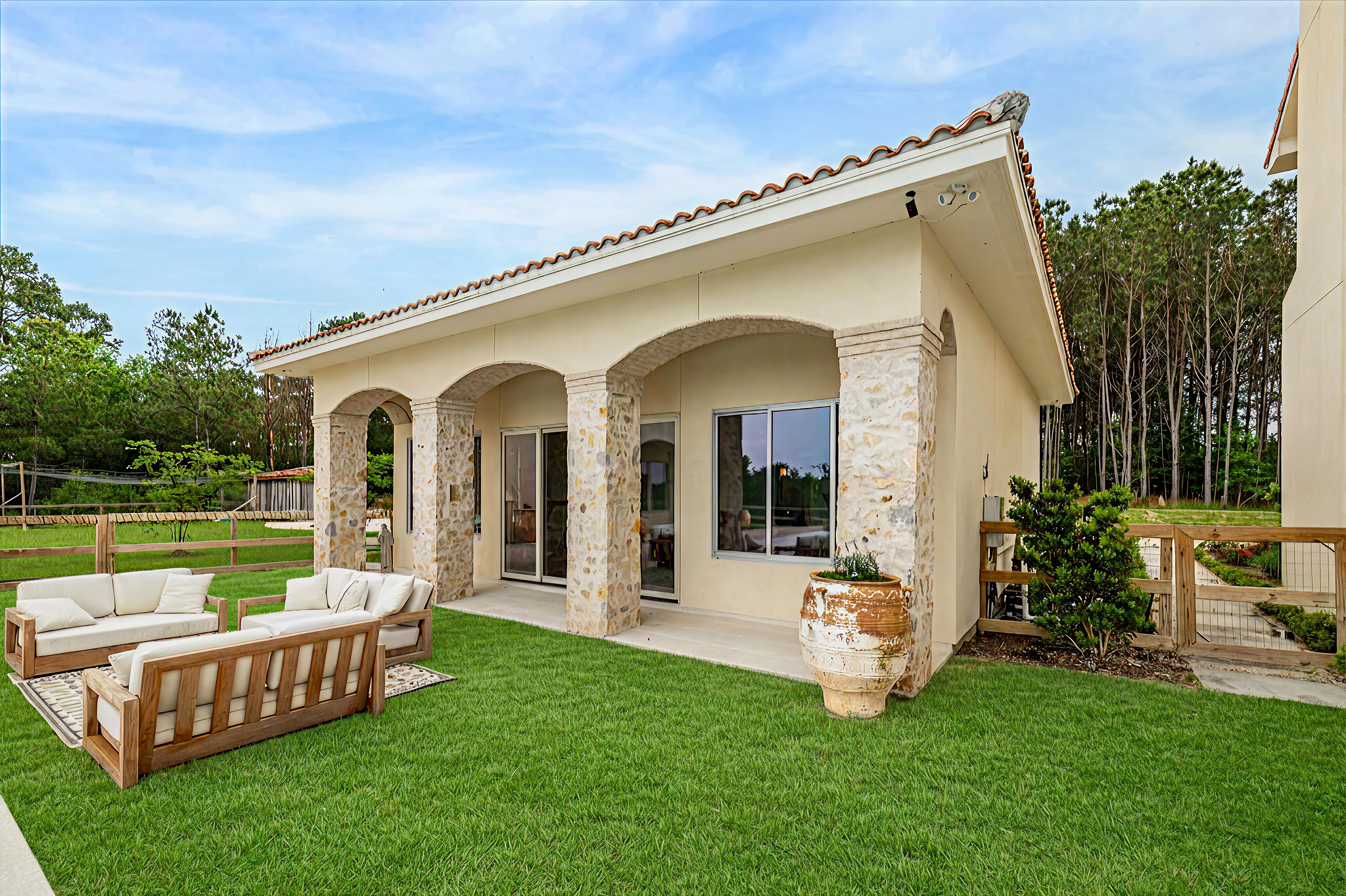 Backyard patio with outdoor furniture, including a sofa, loveseat, and armchair with cream cushions. The patio is attached to a house with stone and stucco exterior, arched openings, and sliding glass doors. The yard has a green lawn, a large ceramic pot, and a wooden fence with trees in the background.