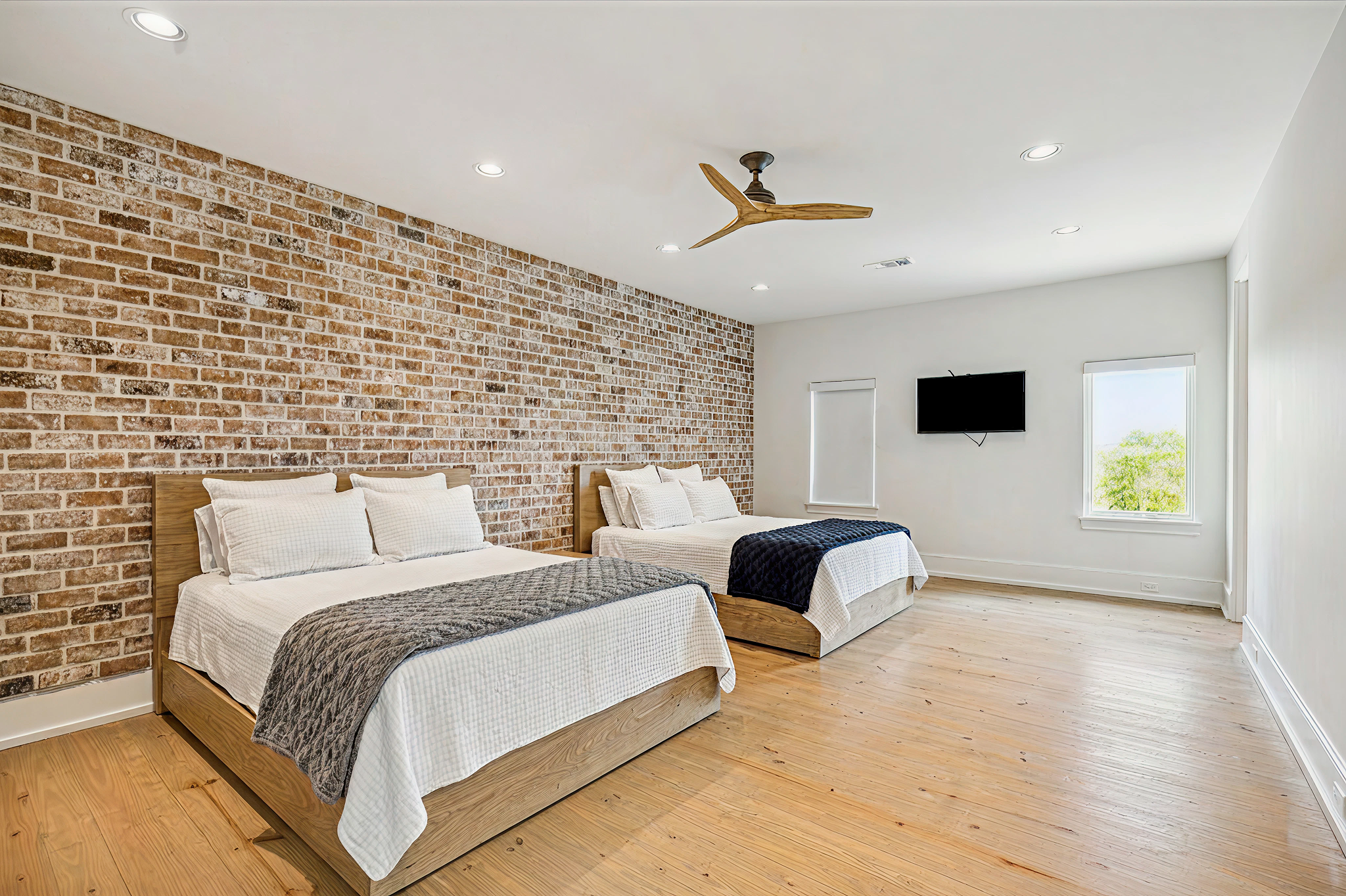 A bedroom with two beds with wood frames, a brick accent wall, a ceiling fan designed like a branch, three recessed ceiling lights, a wall-mounted TV, two windows with white blinds, and light wood flooring.