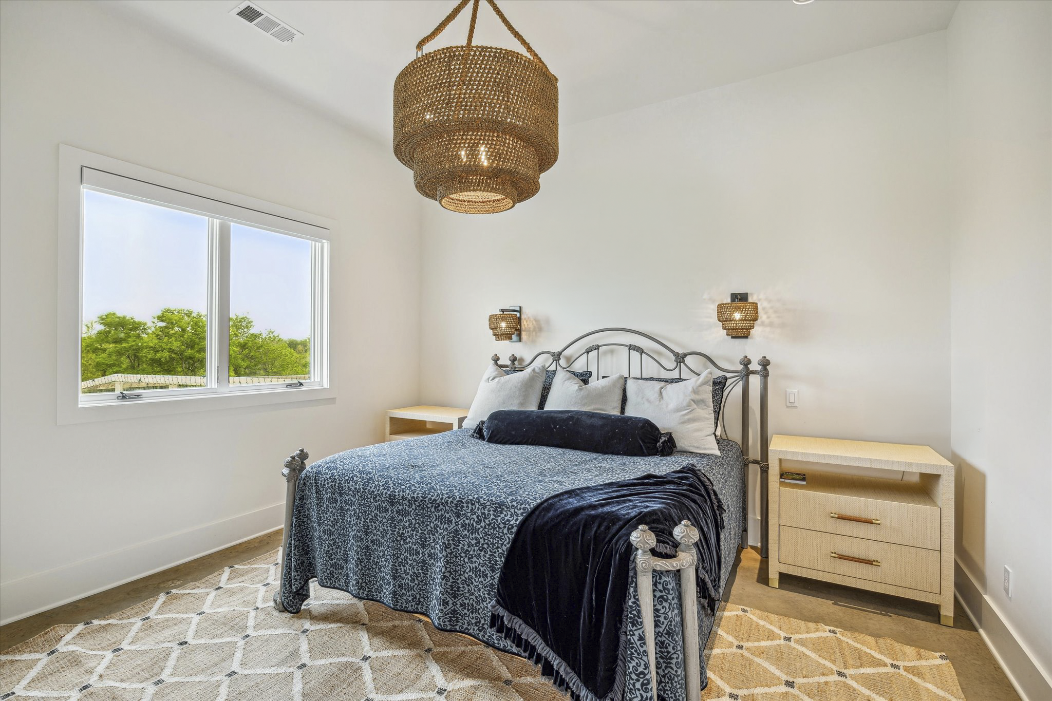 A bedroom with a metal bed frame, navy and white bedding, two side tables, and a woven pendant light hanging from the ceiling. A window with a view of green trees and a beige patterned rug on the floor.