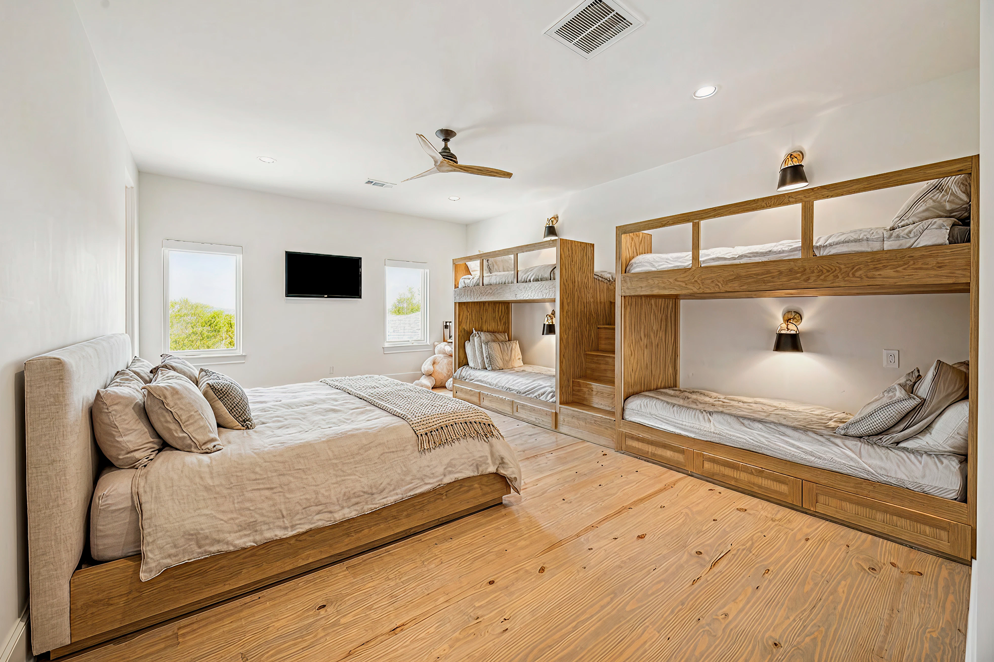 A bedroom with a large bed, two windows, a wall-mounted TV, and custom built-in bunk beds with stairs and reading lights.