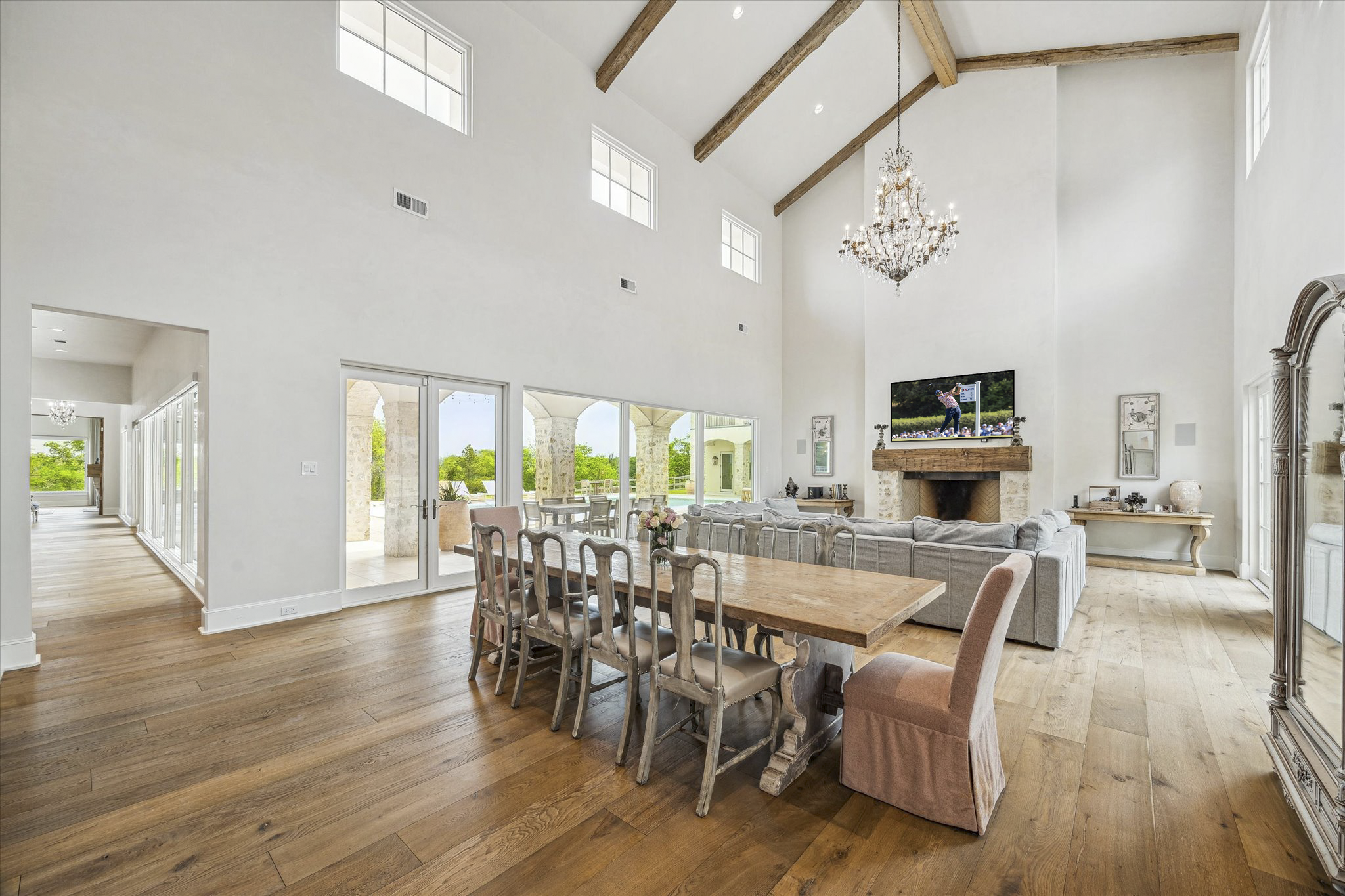 A spacious living and dining room with high white walls, exposed wooden beams on the ceiling, hardwood floors, a chandelier, a large TV above a fireplace, a gray sectional sofa, a wooden dining table with matching chairs, and a sliding glass door leading to an outdoor patio with stone arches and greenery.