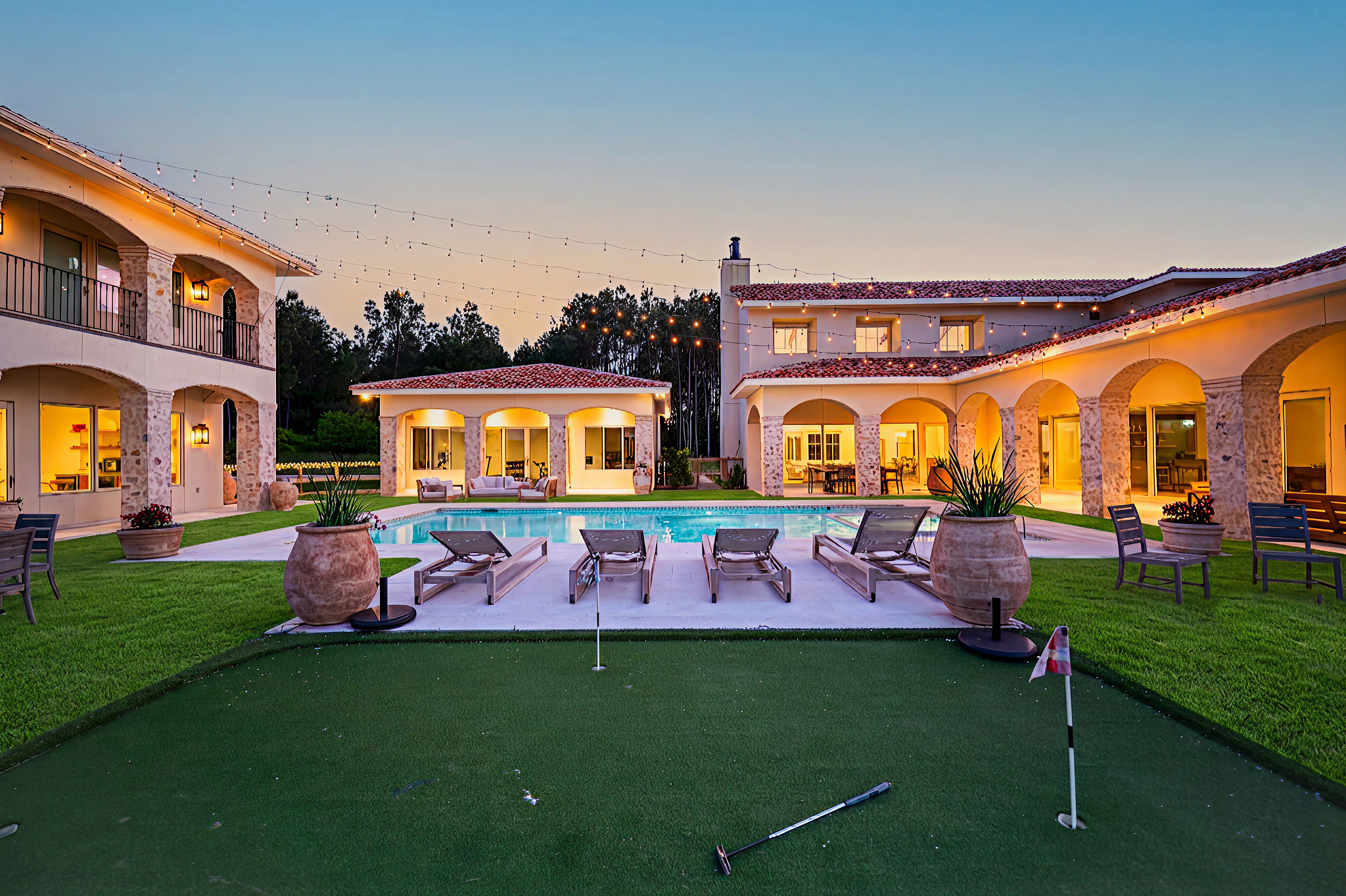 A luxurious outdoor pool area at sunset with lounge chairs, a putting green, and surrounding apartment buildings with string lights and illuminated windows.