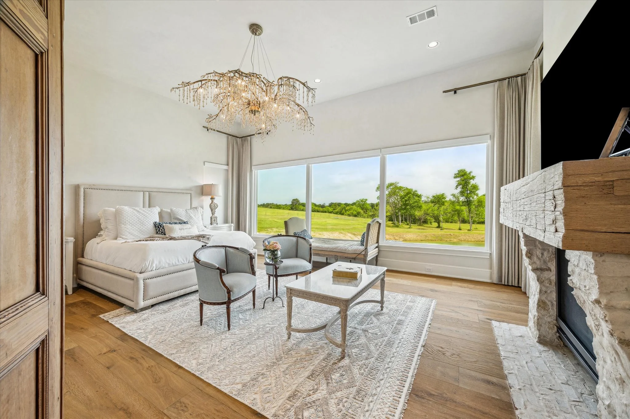 Bright bedroom with large window overlooking green landscape, white sofa, custom chairs, coffee table, chandelier, and stone fireplace.
