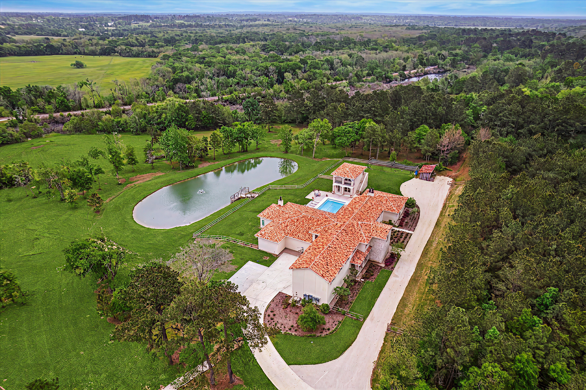 Aerial view of large estate with white house featuring red tile roof, swimming pool, pond, landscaped yard, dirt driveway, and surrounding green trees and fields.