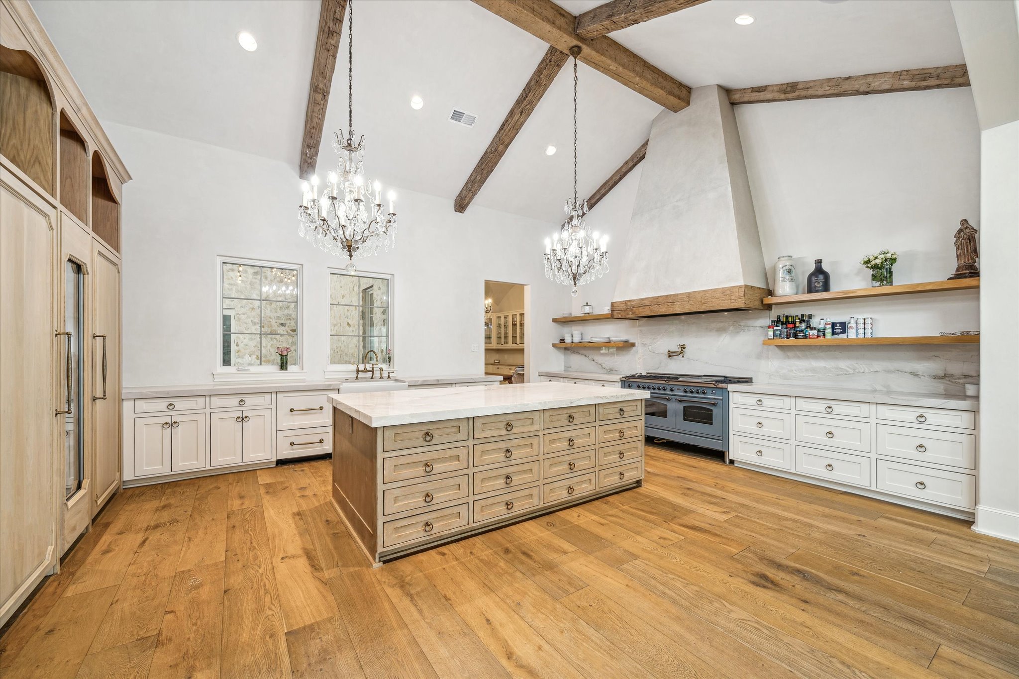 A spacious kitchen with white cabinetry, a large island with drawers, wooden ceiling beams, two chandeliers, a stone wall, open wooden shelves with decorative items, and a blue stove.