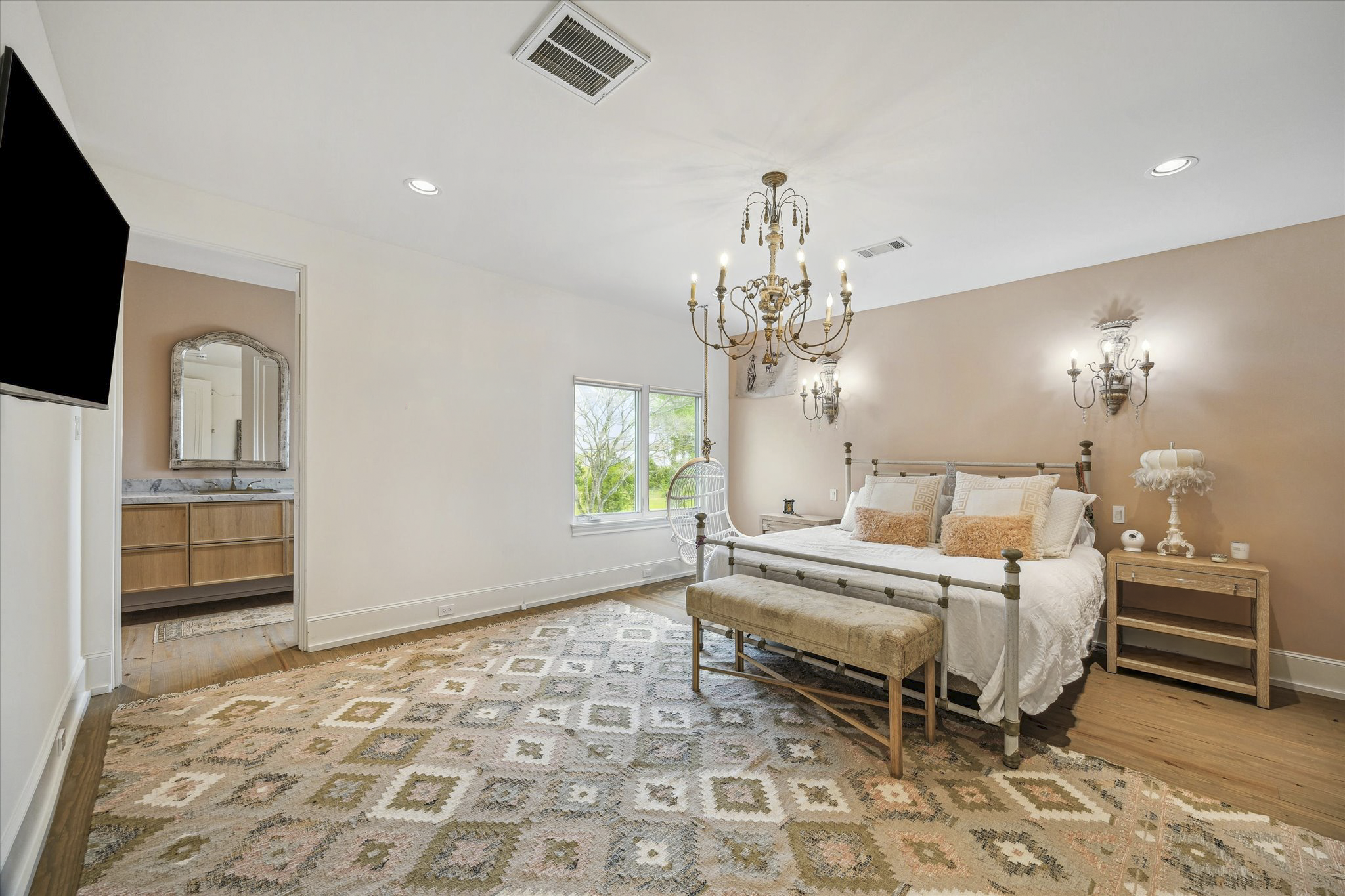 A spacious bedroom with a large bed, nightstands, and decorative wall sconces. The room has a chandelier, a window with a view of trees outside, and a patterned area rug on wooden flooring. An entryway reveals an adjoining bathroom with a mirror and vanity.
