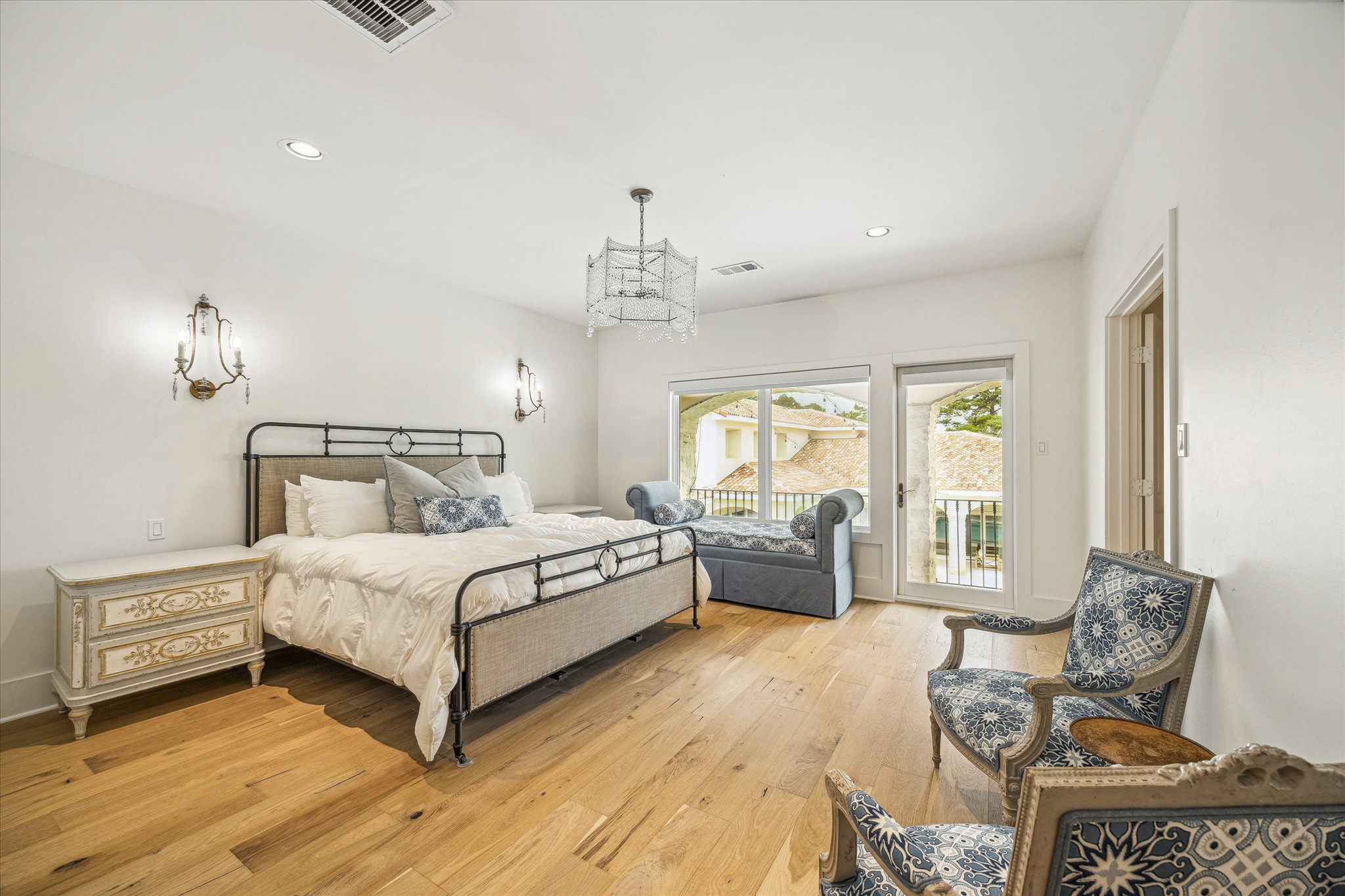 A bedroom with a metal bedframe, white bedding, blue and white decorative pillows, vintage nightstand, and a sitting area with blue upholstered chairs near a window and balcony door, featuring hardwood floors, white walls, and decorative wall sconces.