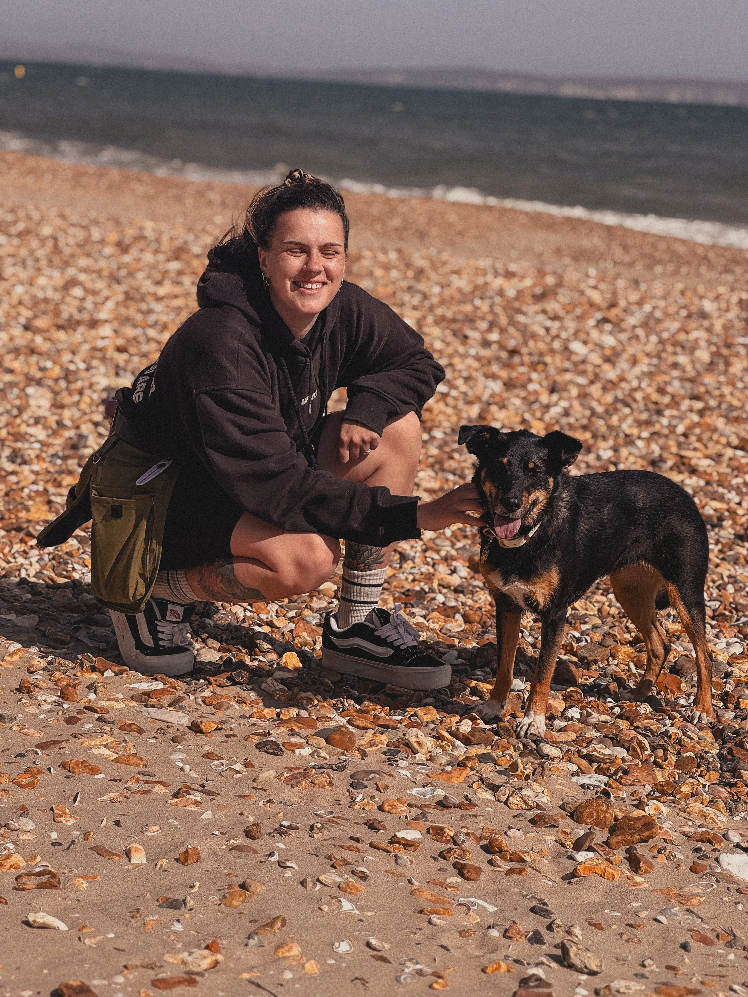 A woman crouching on a pebble beach next to a black and brown dog, smiling as she pets the dog with her right hand, with the ocean and sky in the background.