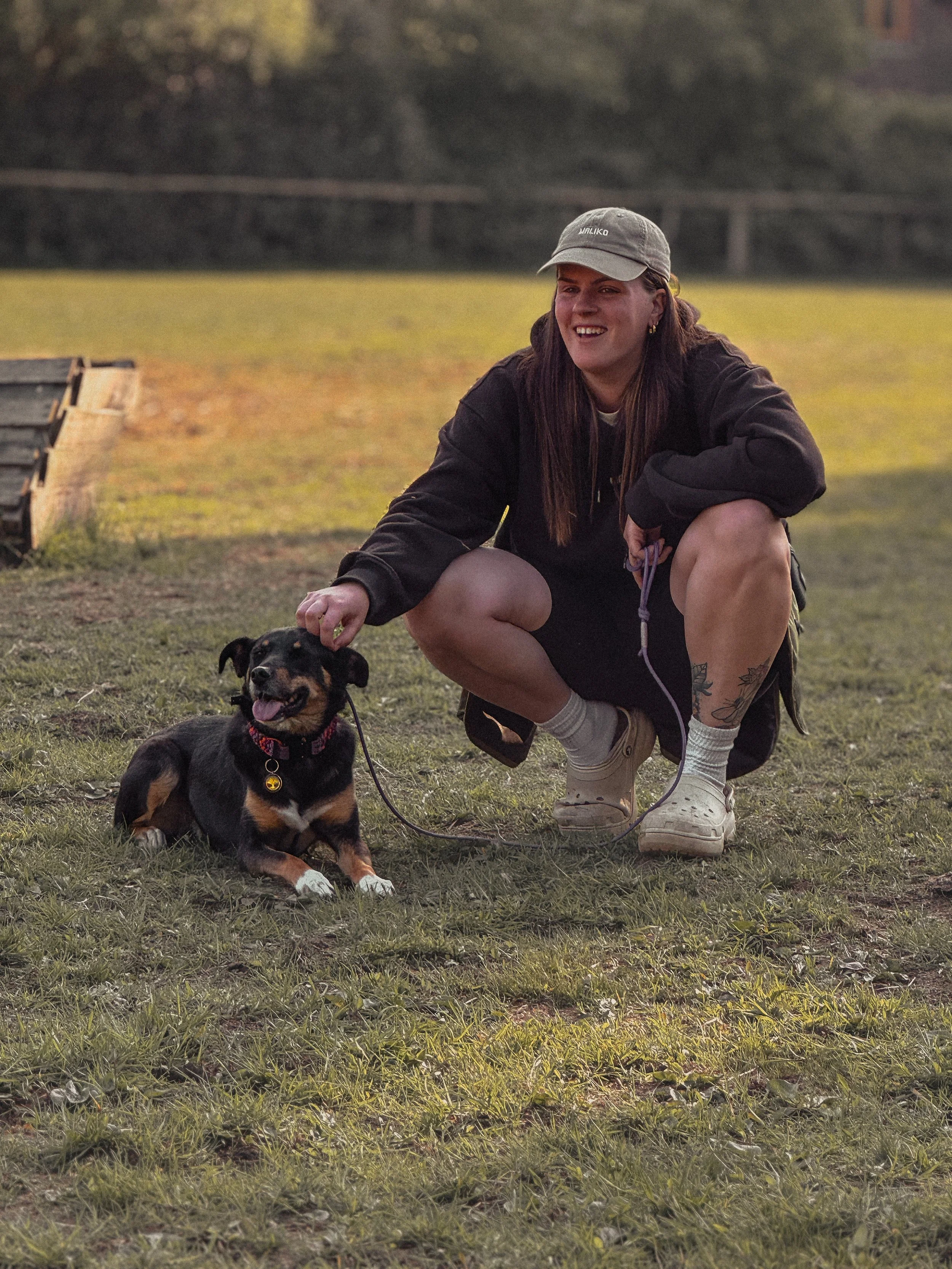 A woman crouching on grass next to a small black and brown dog. The woman is wearing a gray baseball cap, black hoodie, white socks, and white sneakers, smiling and petting the dog. The dog has a red collar and is sitting on the grass with its tongue out.