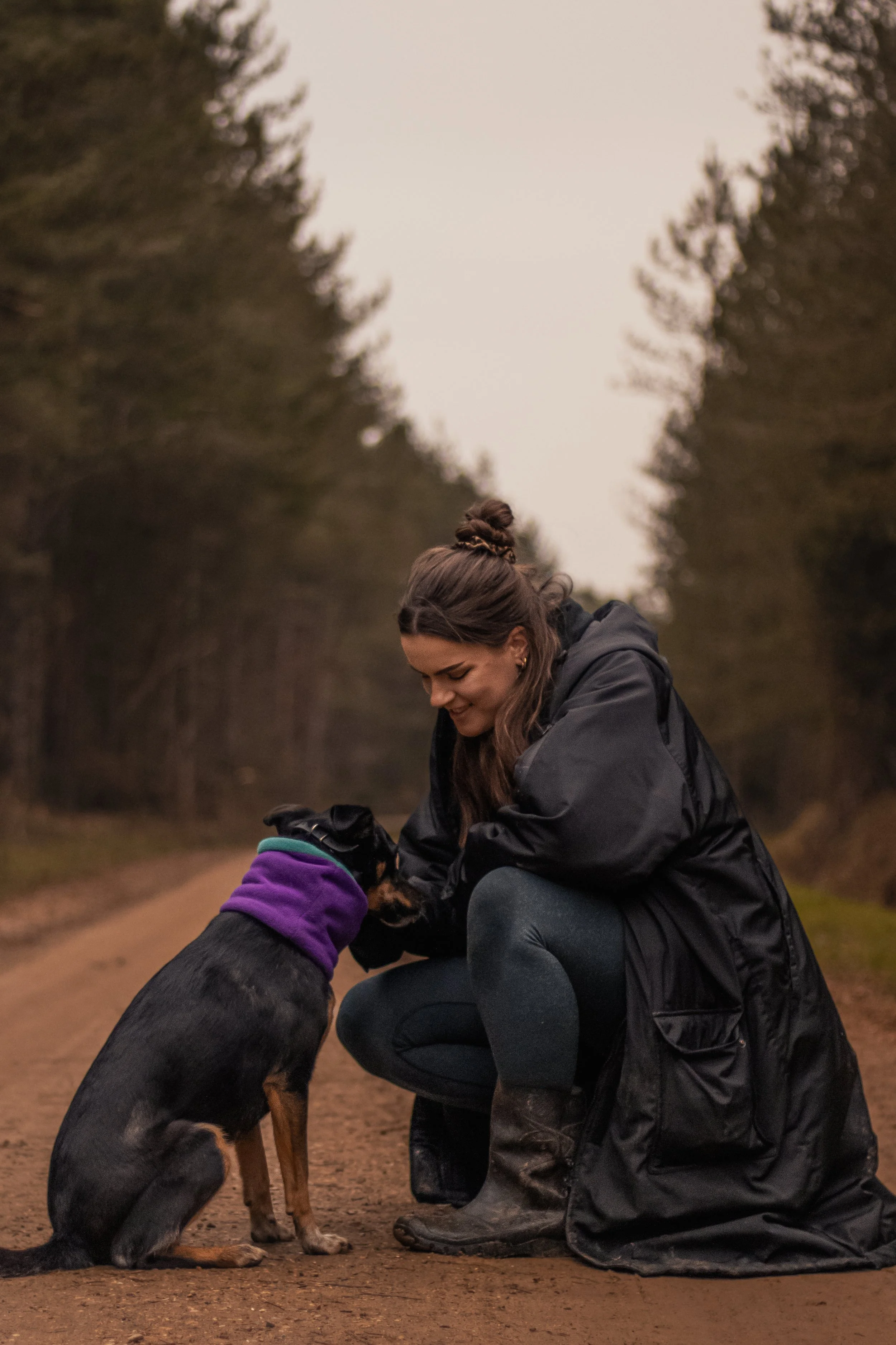 A woman kneeling on a dirt forest road, smiling while petting a black dog wearing a purple scarf and gloves, with trees lining the road in the background.