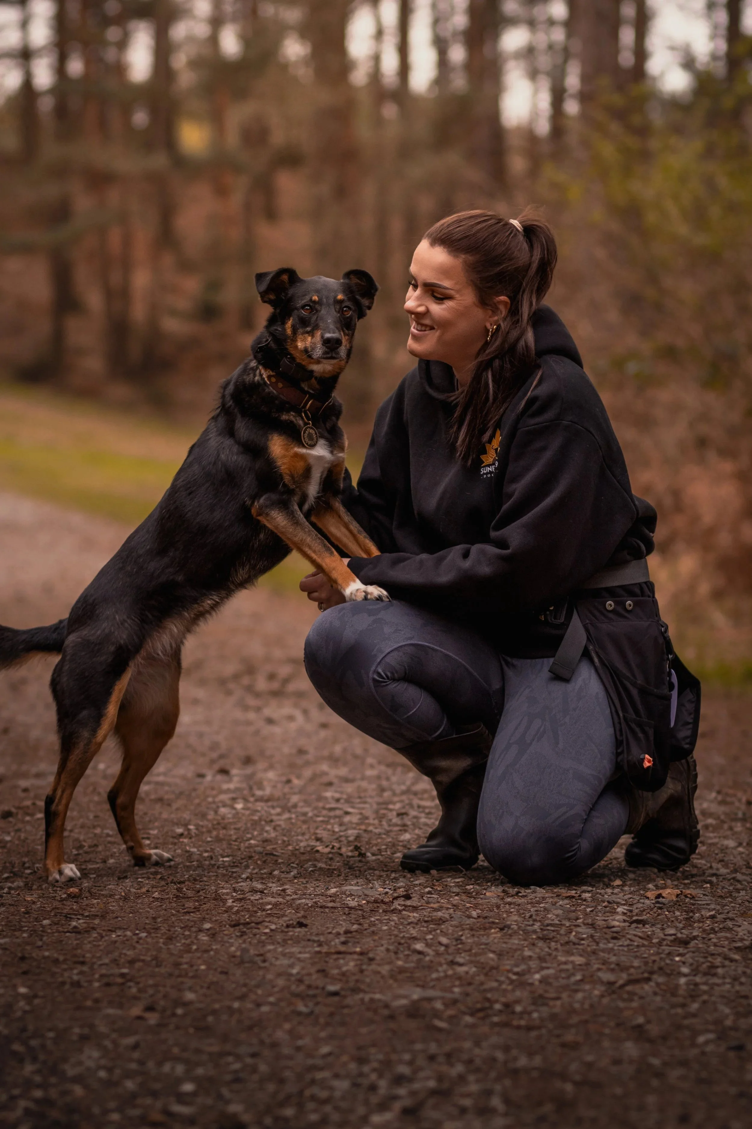 A woman squatting outdoors with a black and brown dog in a wooded area, both appearing happy and engaged.
