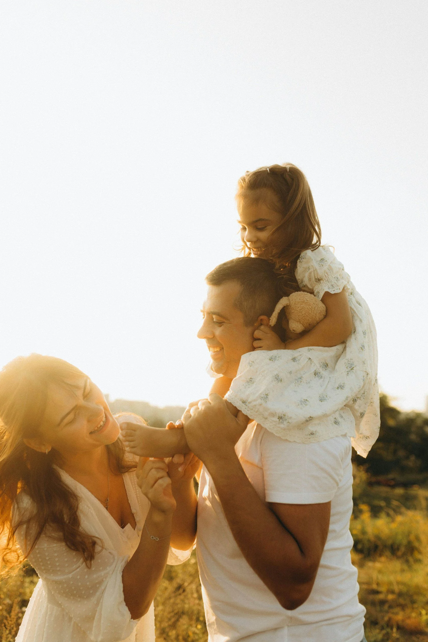 A happy family enjoying a moment outdoors during sunset, with a man carrying a young girl on his shoulders and a woman playing with the girl, all smiling and laughing.